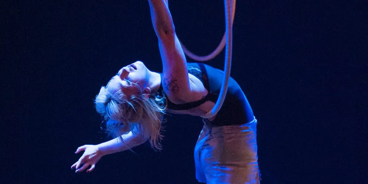 L'artiste aérienne Alyssa Bunce se suspend gracieusement à un cerceau, le dos arqué, les bras tendus et une jambe pointée, éclairée par la lumière bleue de la scène.