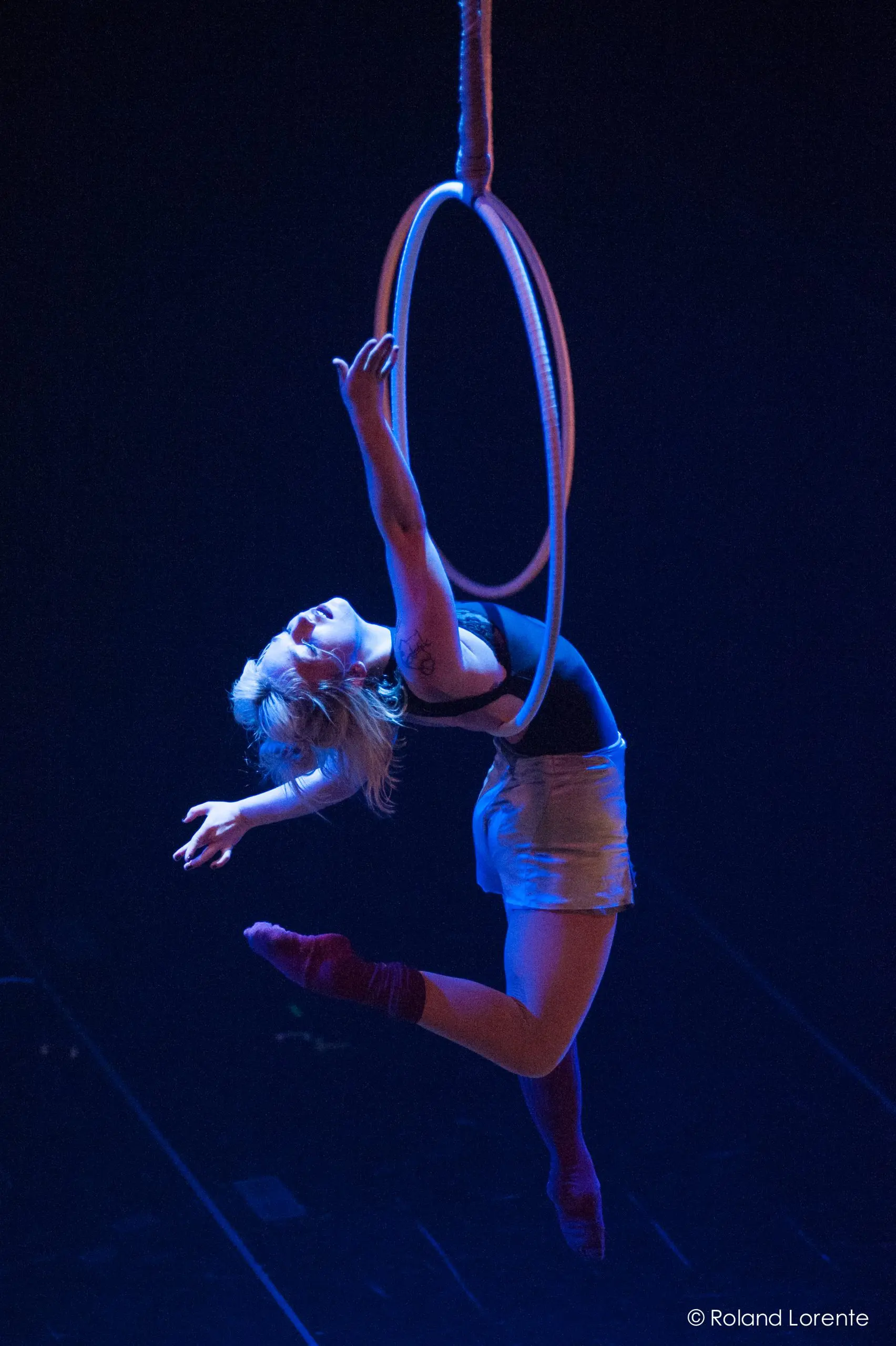 L'artiste aérienne Alyssa Bunce se suspend gracieusement à un cerceau, le dos arqué, les bras tendus et une jambe pointée, éclairée par la lumière bleue de la scène.