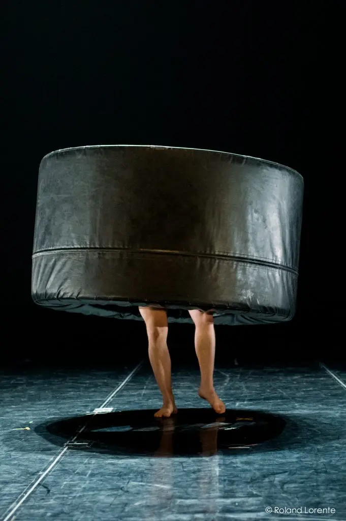 Une paire de jambes nues s'étend sous un grand objet rond et noir rembourré sur une scène sombre, créant une scène surréaliste et ludique qui rappelle l'art fantaisiste de Basile Philippe.