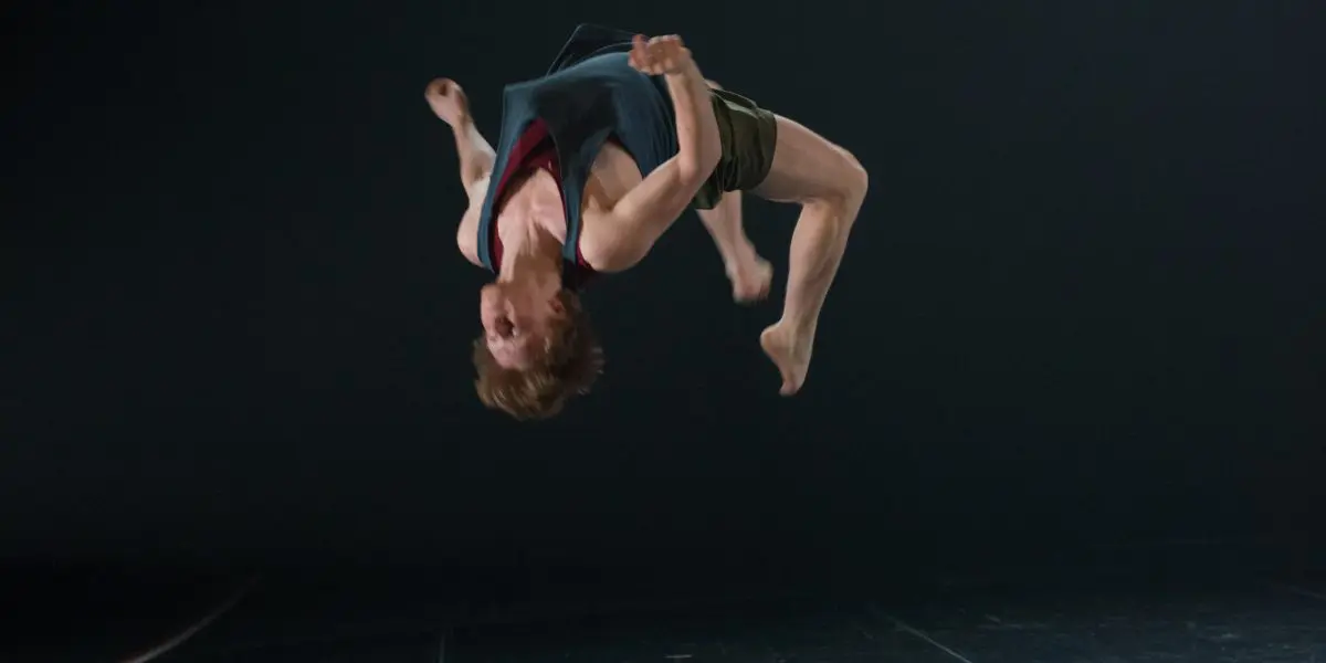 Un danseur exécute un saut périlleux arrière sur une scène faiblement éclairée, tandis qu'une autre personne est allongée sur le sol, partiellement éclairée par les lumières de la scène dans cette scène hypnotique chorégraphiée par Basile Philippe.