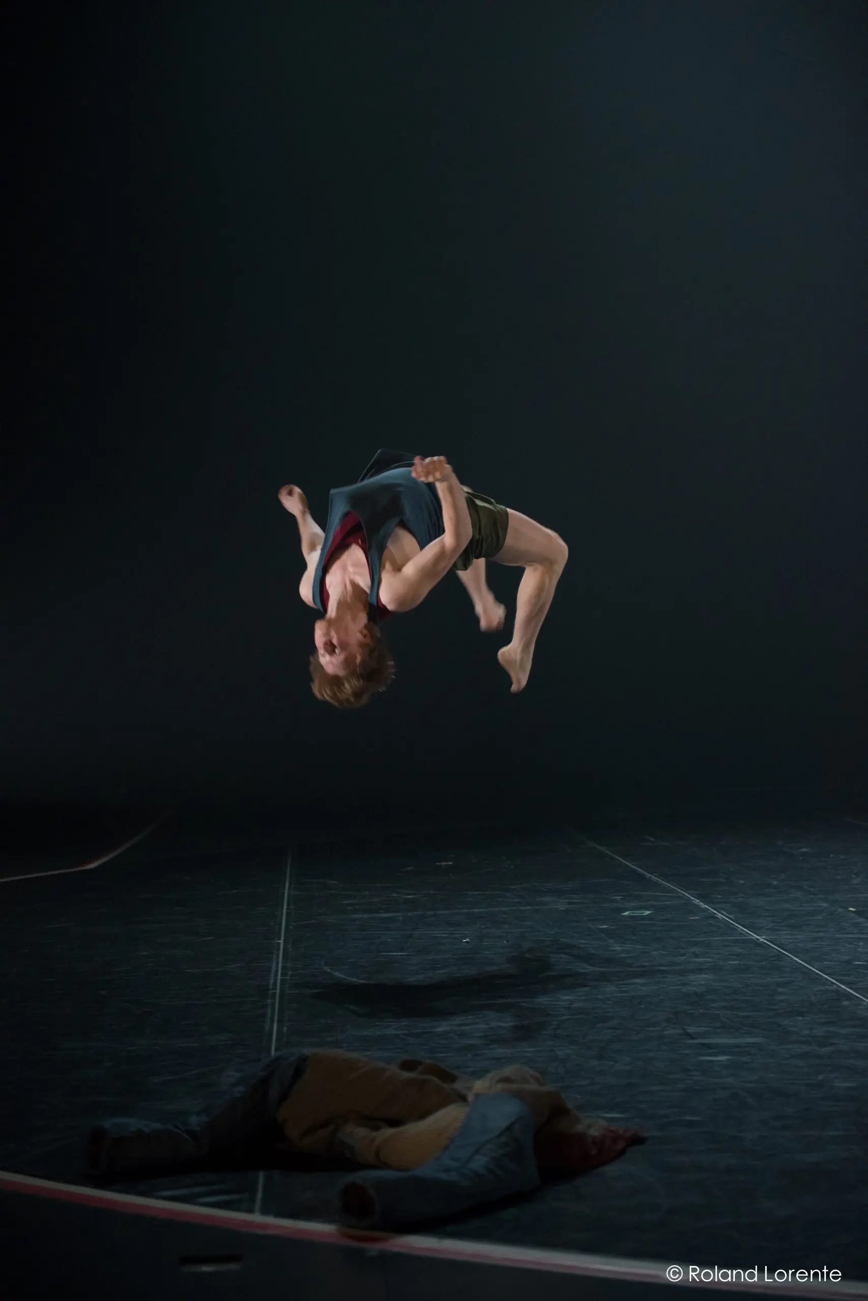 Un danseur exécute un saut périlleux arrière sur une scène faiblement éclairée, tandis qu'une autre personne est allongée sur le sol, partiellement éclairée par les lumières de la scène dans cette scène hypnotique chorégraphiée par Basile Philippe.