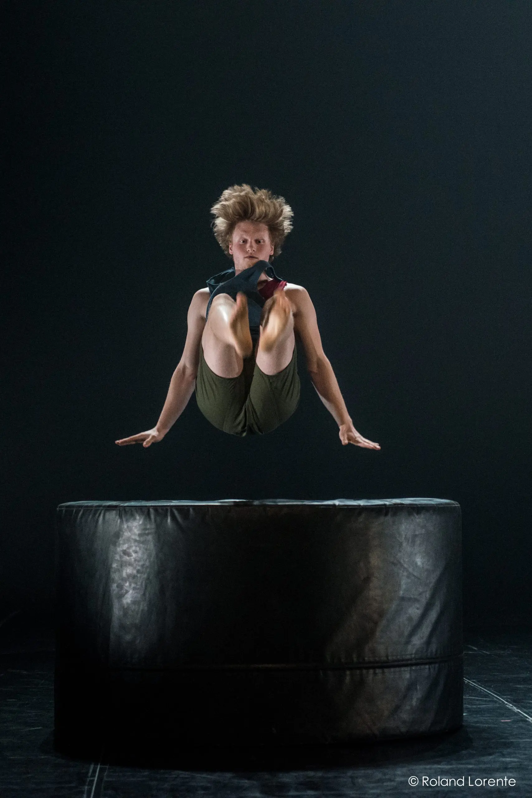 Une personne aux cheveux ébouriffés est capturée en plein vol, les genoux ramenés à la poitrine, au-dessus d'une grande plate-forme ronde et rembourrée, effectuant un saut acrobatique dans un espace faiblement éclairé, rappelant les photographies de mouvements dynamiques de Basile Philippe.