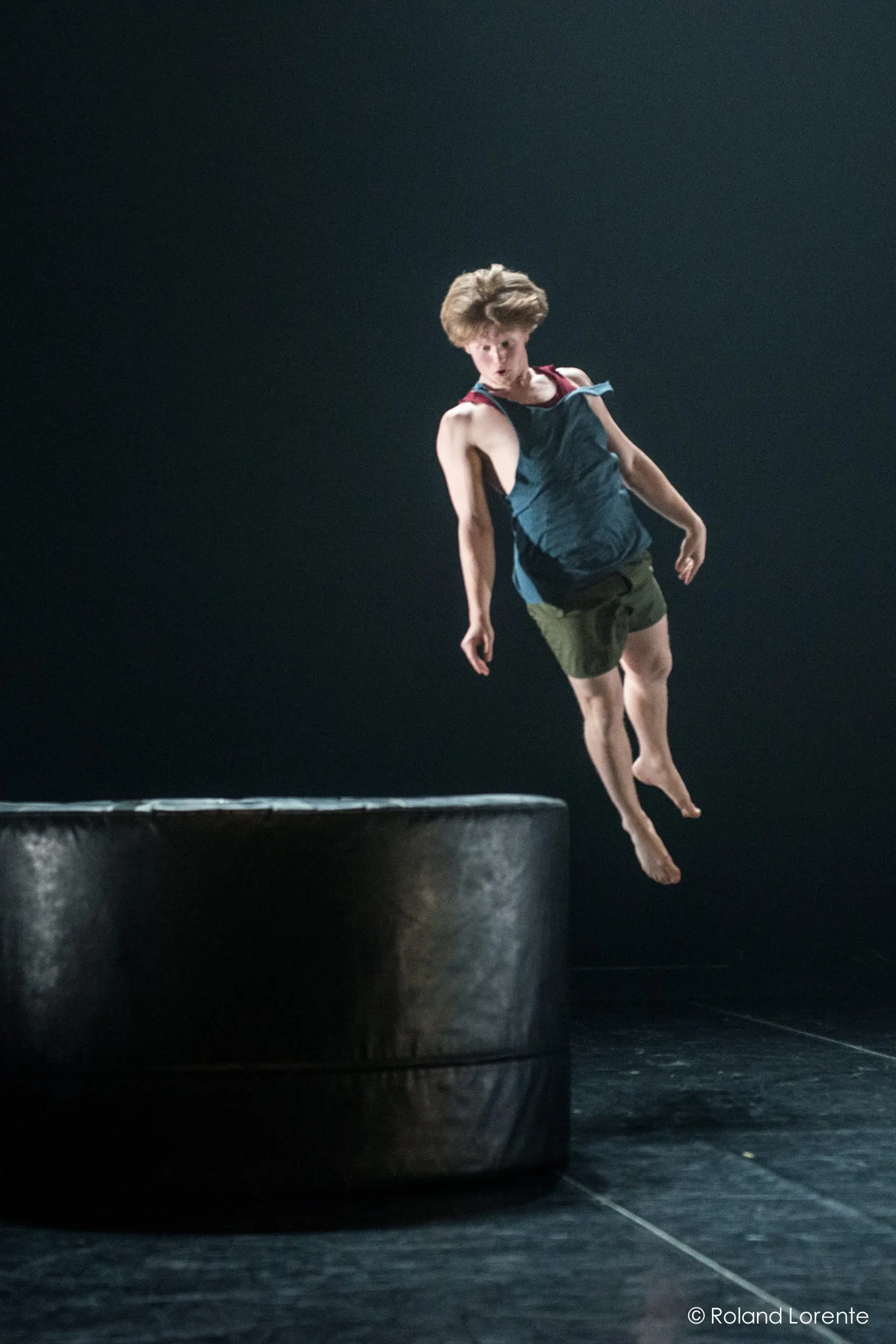 Un jeune homme vêtu d'un débardeur et d'un short est capturé en plein vol, semblant sauter ou tomber près d'une grande plate-forme ronde et rembourrée de couleur noire sur une scène faiblement éclairée - un moment dynamique qui rappelle les performances de Basile Philippe.