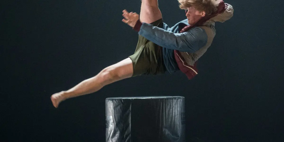 Un danseur est capturé dans les airs au-dessus d'une plate-forme cylindrique noire, une jambe tendue vers le haut et les bras tendus, se produisant sous un projecteur sur une scène sombre - un moment qui rappelle le style dynamique de Basile Philippe.