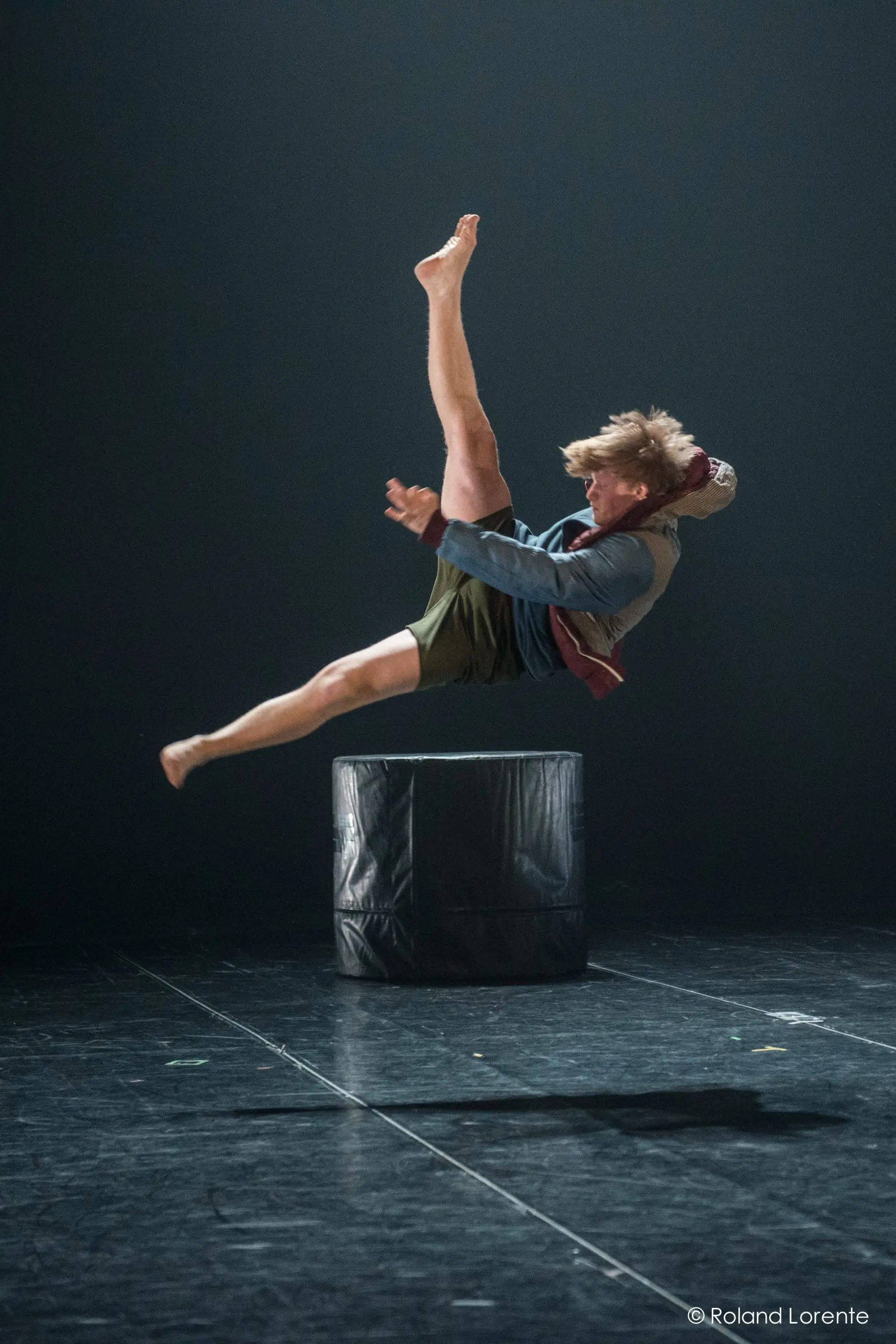 Un danseur est capturé dans les airs au-dessus d'une plate-forme cylindrique noire, une jambe tendue vers le haut et les bras tendus, se produisant sous un projecteur sur une scène sombre - un moment qui rappelle le style dynamique de Basile Philippe.
