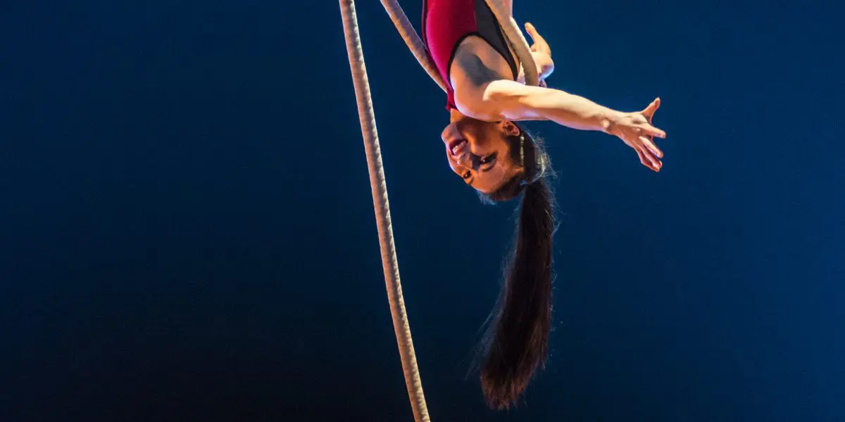 Une voltigeuse, Brittany Gee-Moore, vêtue d'un justaucorps rouge, prend la pose à l'envers sur une corde épaisse, étendant un bras et souriant sous un éclairage dramatique sur un fond bleu foncé.