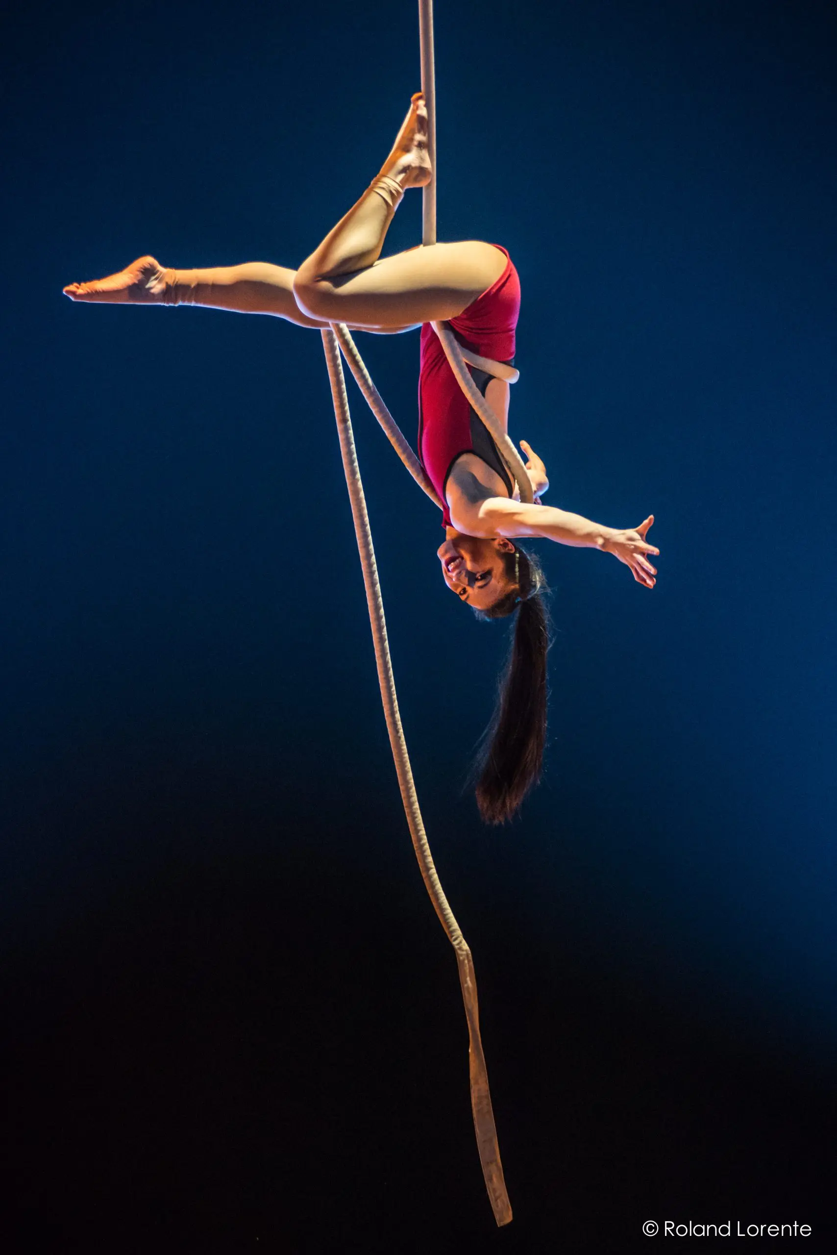 Une voltigeuse, Brittany Gee-Moore, vêtue d'un justaucorps rouge, prend la pose à l'envers sur une corde épaisse, étendant un bras et souriant sous un éclairage dramatique sur un fond bleu foncé.