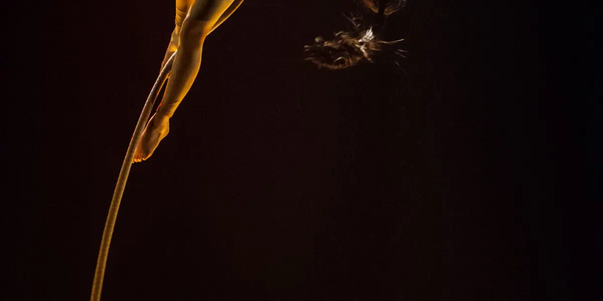 L'artiste aérienne Brittany Gee-Moore, vêtue d'un justaucorps rouge, est suspendue à une corde, la tête en bas, le dos arqué, un bras tendu et les cheveux flottants, sur un fond sombre.