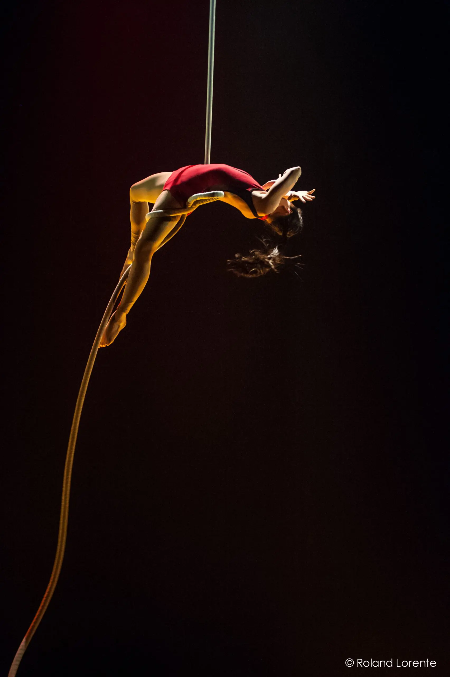 L'artiste aérienne Brittany Gee-Moore, vêtue d'un justaucorps rouge, est suspendue à une corde, la tête en bas, le dos arqué, un bras tendu et les cheveux flottants, sur un fond sombre.