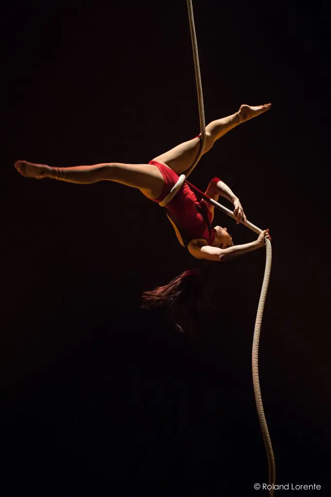 Une artiste aérienne, Brittany Gee-Moore, vêtue d'une tenue rouge, est suspendue à l'envers à une corde, les jambes étendues en fente, sur un fond sombre.