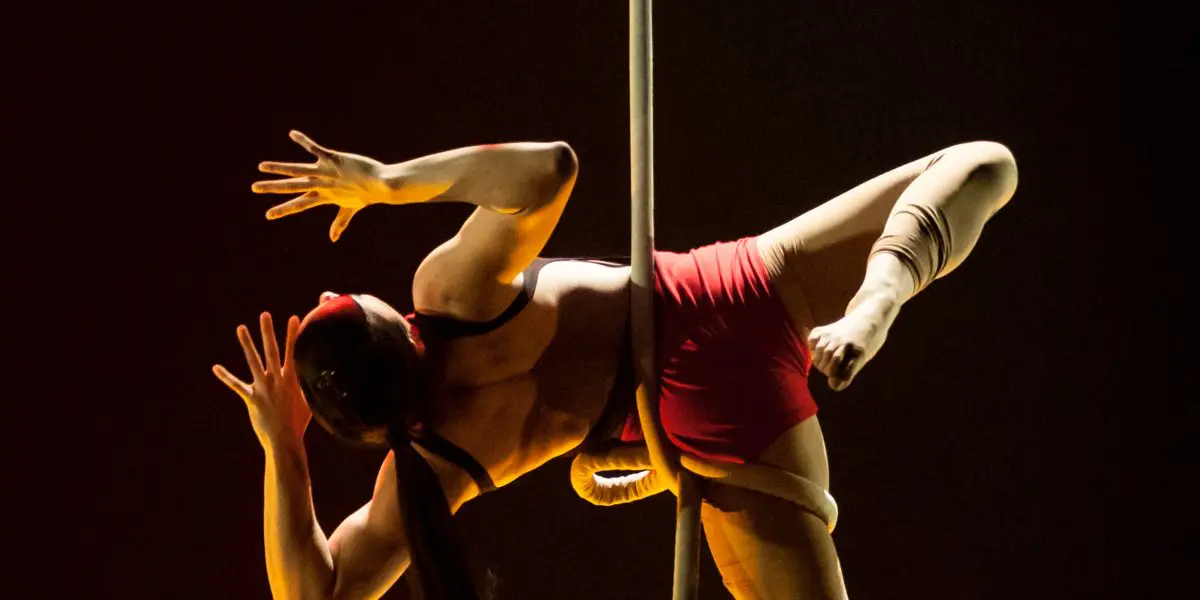 Une acrobate, Brittany Gee-Moore, vêtue d'un short rouge et d'une longue queue de cheval, effectue une pose aérienne sur un poteau vertical, éclairée par un éclairage de scène spectaculaire sur un fond sombre.