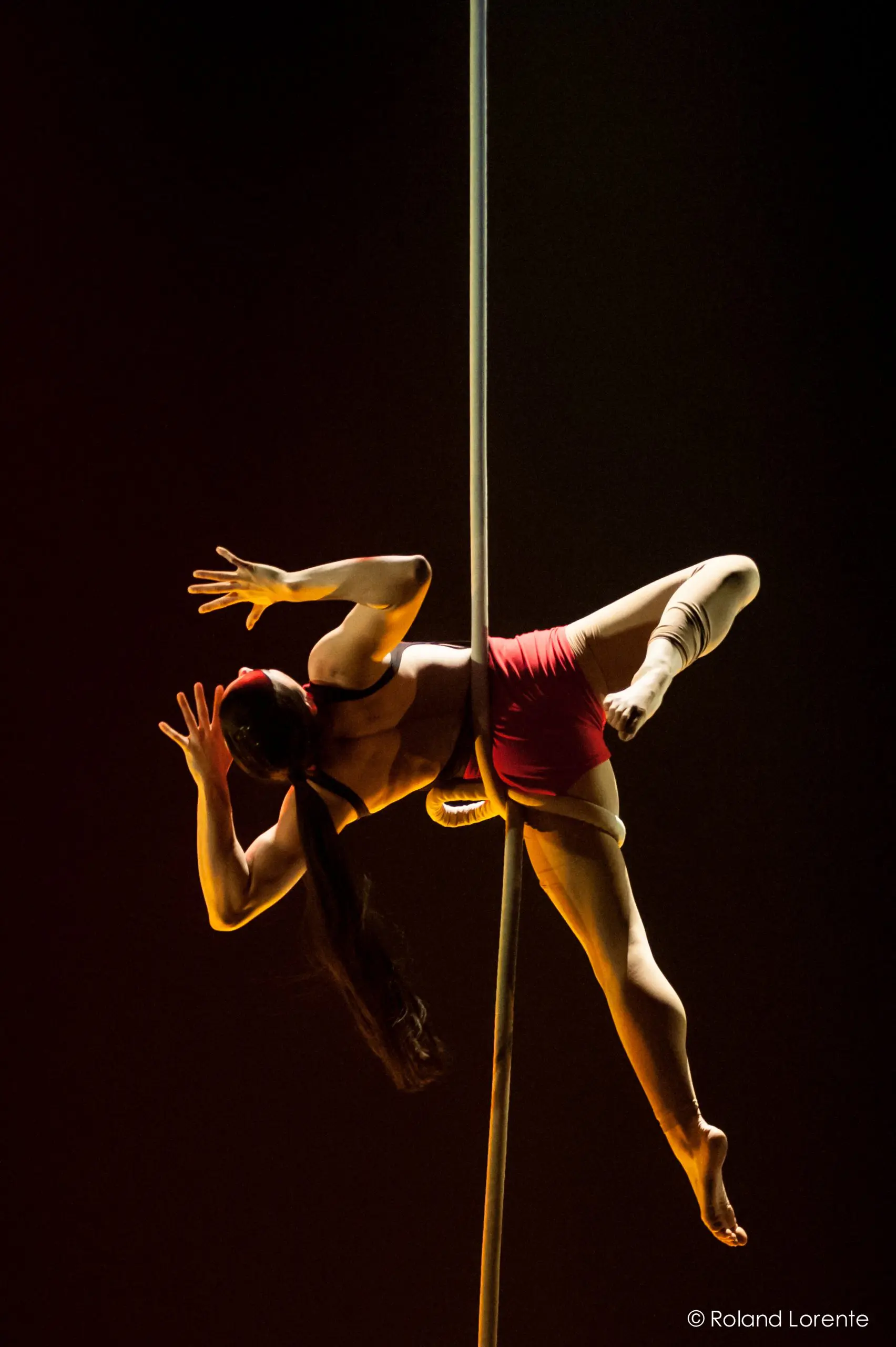 Une acrobate, Brittany Gee-Moore, vêtue d'un short rouge et d'une longue queue de cheval, effectue une pose aérienne sur un poteau vertical, éclairée par un éclairage de scène spectaculaire sur un fond sombre.