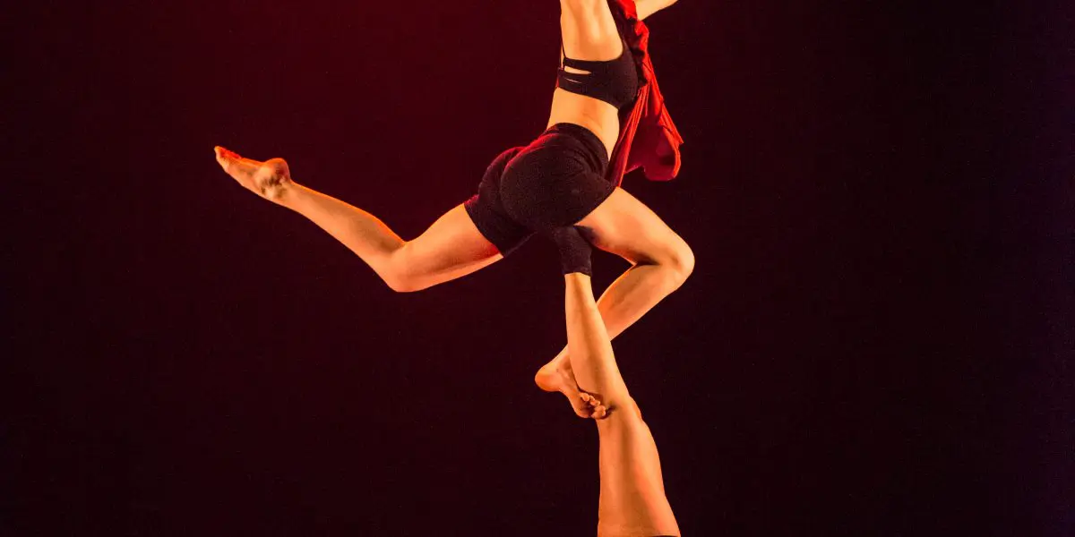 Deux acrobates exécutent un numéro d'équilibre ; Catherine Beaudet se tient sur les mains tandis que l'autre se tient en équilibre sur ses pieds, une jambe tendue et un bras levé, tous deux dramatiquement éclairés par un éclairage de scène rouge.
