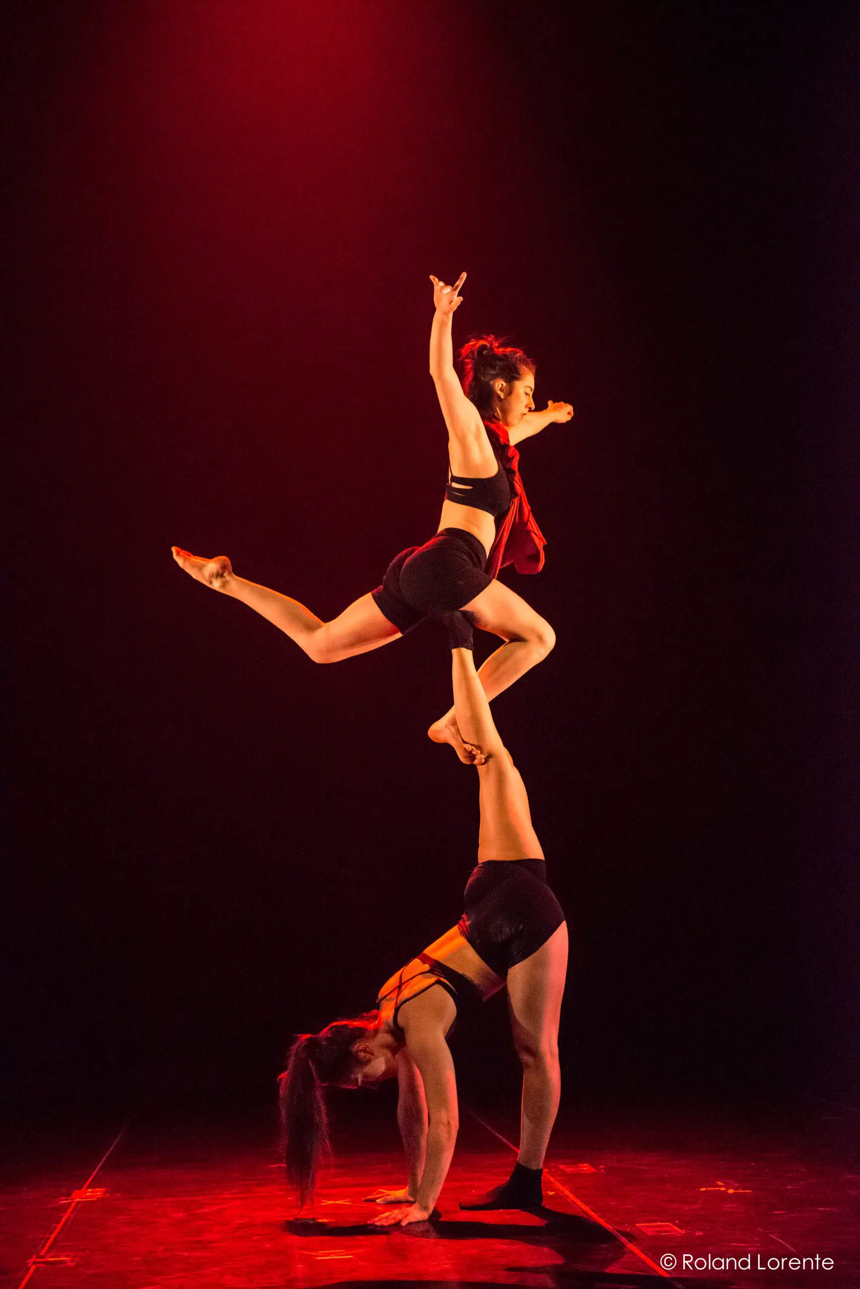 Deux acrobates exécutent un numéro d'équilibre ; Catherine Beaudet se tient sur les mains tandis que l'autre se tient en équilibre sur ses pieds, une jambe tendue et un bras levé, tous deux dramatiquement éclairés par un éclairage de scène rouge.