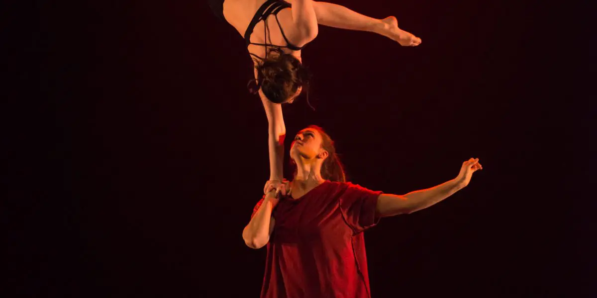 Deux acrobates évoluent sur scène sous un éclairage rouge ; Catherine Beaudet tient son partenaire en équilibre sur son bras, la tête en bas, faisant preuve d'une force et d'un équilibre remarquables.