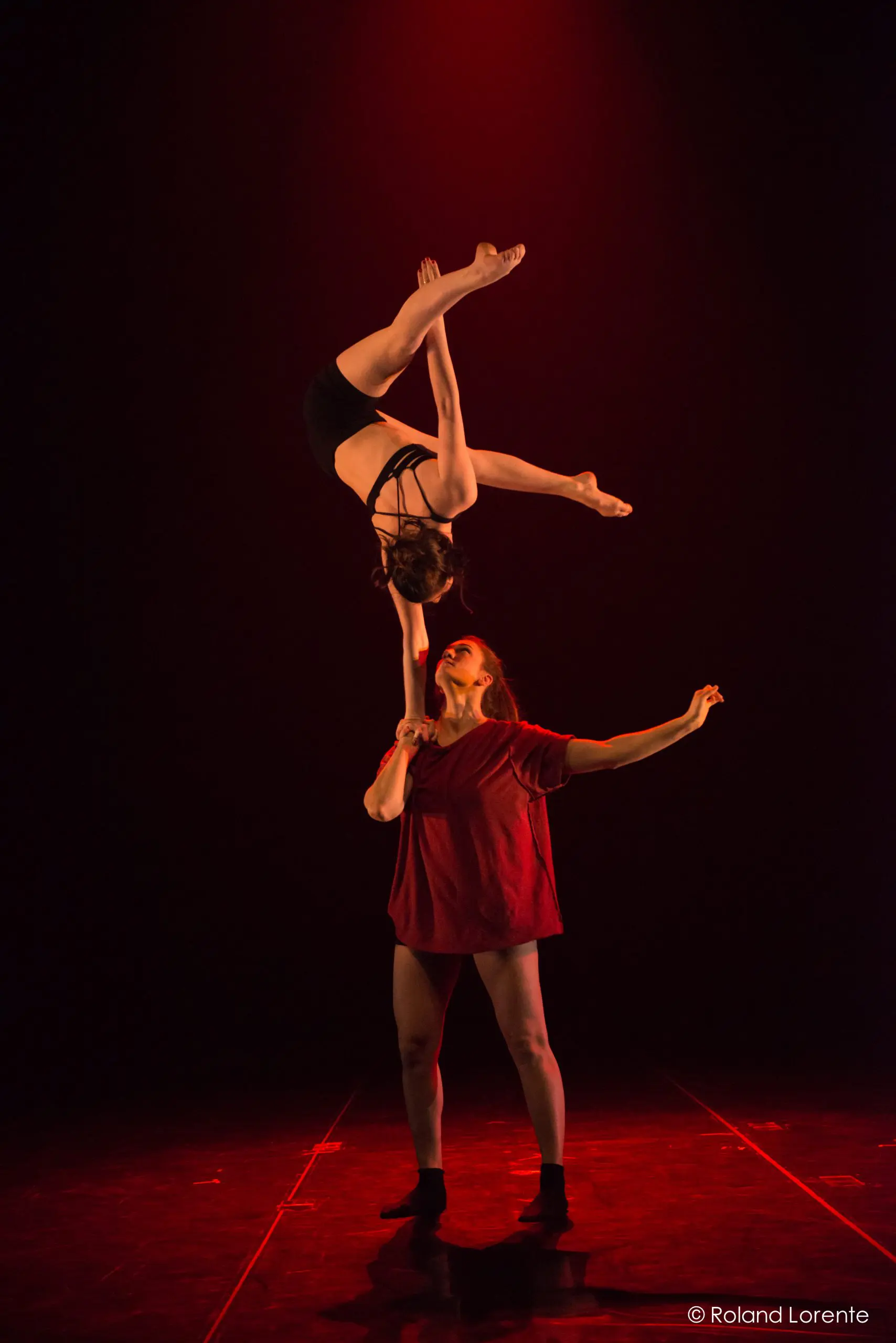 Deux acrobates évoluent sur scène sous un éclairage rouge ; Catherine Beaudet tient son partenaire en équilibre sur son bras, la tête en bas, faisant preuve d'une force et d'un équilibre remarquables.