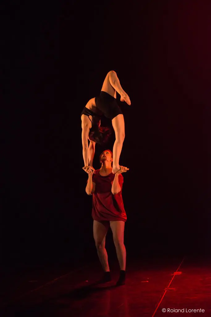 Deux acrobates évoluent sur une scène sombre. Catherine Beaudet est en équilibre sur les mains de son partenaire, les jambes repliées au-dessus de sa tête, tandis que le socle se tient fermement, éclairé par une lumière rouge.