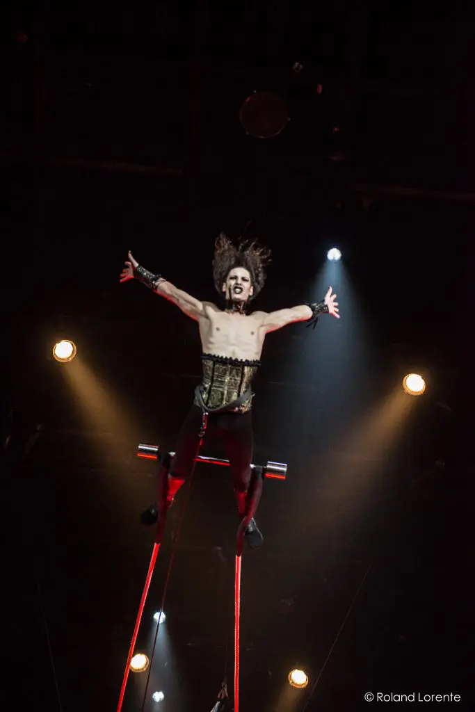 Une artiste de cirque vêtue d'un corset, de jambières et de brassards est suspendue dans les airs au-dessus d'un trapèze, les bras tendus, sous un éclairage dramatique - capturant l'art et le flair de Célien Pinon sous les lumières éblouissantes de la scène.