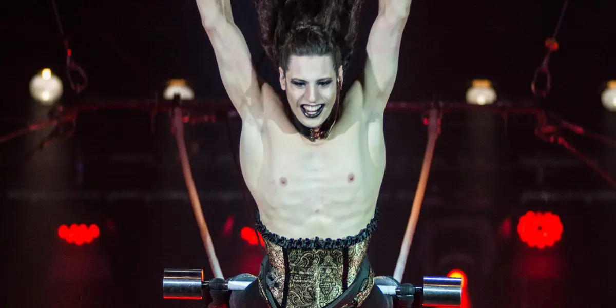 Une artiste de cirque aux cheveux longs, au maquillage spectaculaire et au costume de style corset est en plein vol, les bras levés, semblant sauter ou voler au-dessus d'un trapèze sous un éclairage de scène théâtral dans le style de Célien Pinon.
