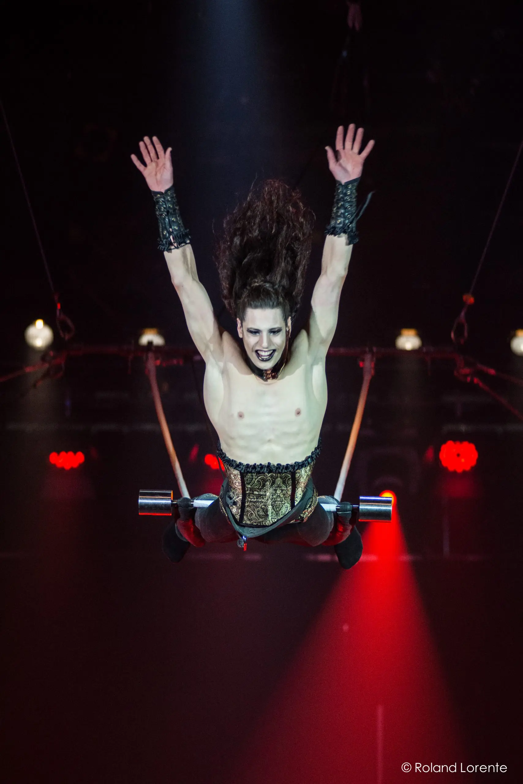 Une artiste de cirque aux cheveux longs, au maquillage spectaculaire et au costume de style corset est en plein vol, les bras levés, semblant sauter ou voler au-dessus d'un trapèze sous un éclairage de scène théâtral dans le style de Célien Pinon.