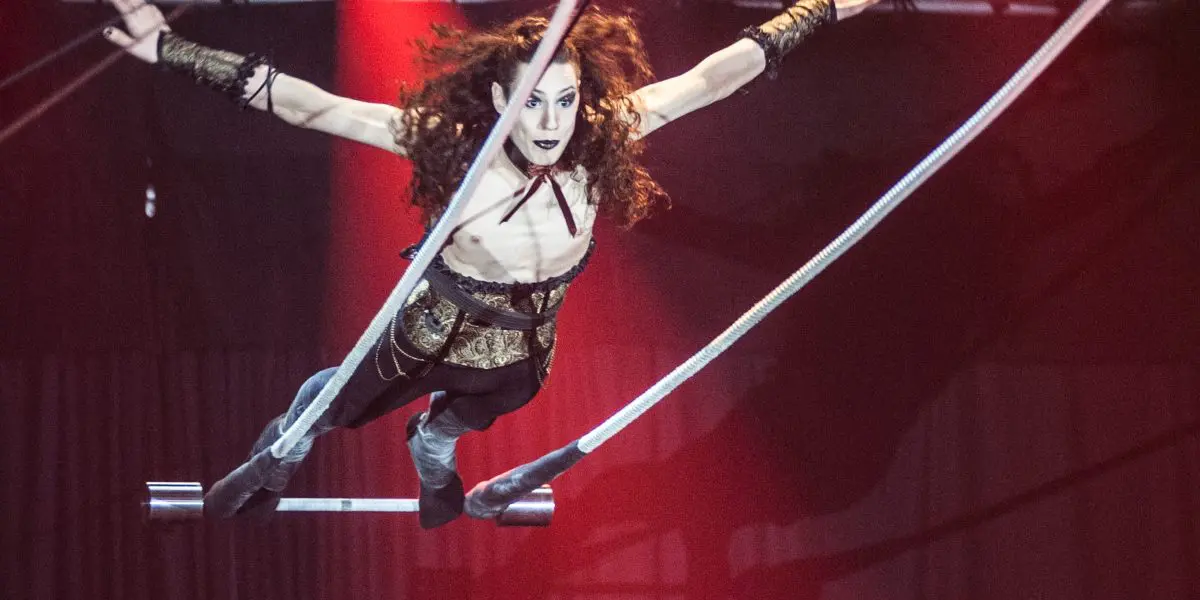 Une trapéziste aux longs cheveux bouclés évolue dans les airs, bras tendus, sous les lumières de la scène et devant les rideaux rouges - un moment captivant qui rappelle le spectaculaire art du cirque de Célien Pinon.