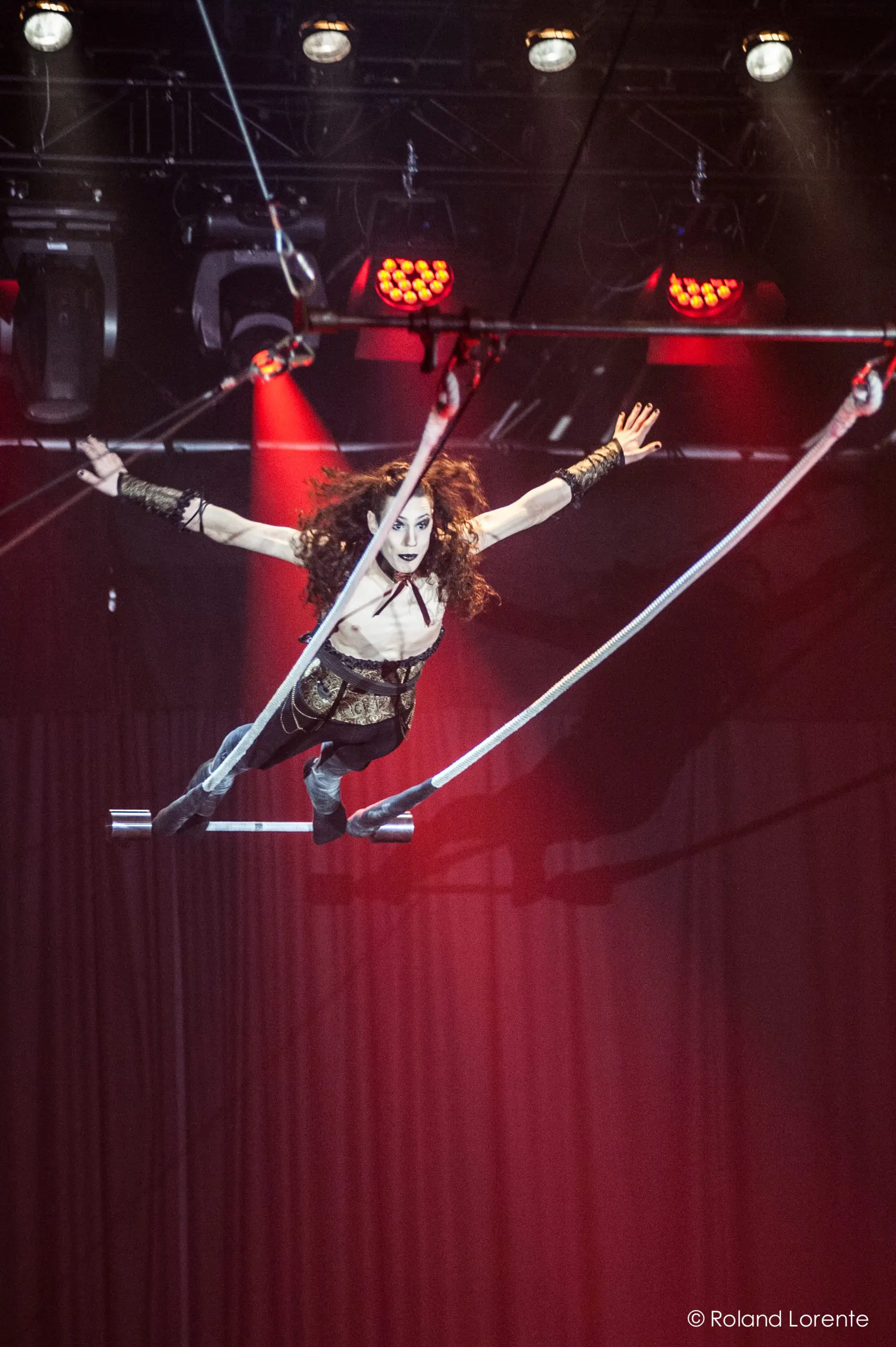 Une trapéziste aux longs cheveux bouclés évolue dans les airs, bras tendus, sous les lumières de la scène et devant les rideaux rouges - un moment captivant qui rappelle le spectaculaire art du cirque de Célien Pinon.