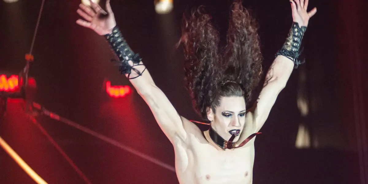 Une artiste de cirque aux cheveux longs, au maquillage spectaculaire et au corset métallique est suspendue dans les airs par des fils - un numéro à couper le souffle qui rappelle l'art de Célien Pinon - les bras tendus sur un fond sombre avec des lumières de scène rouges.