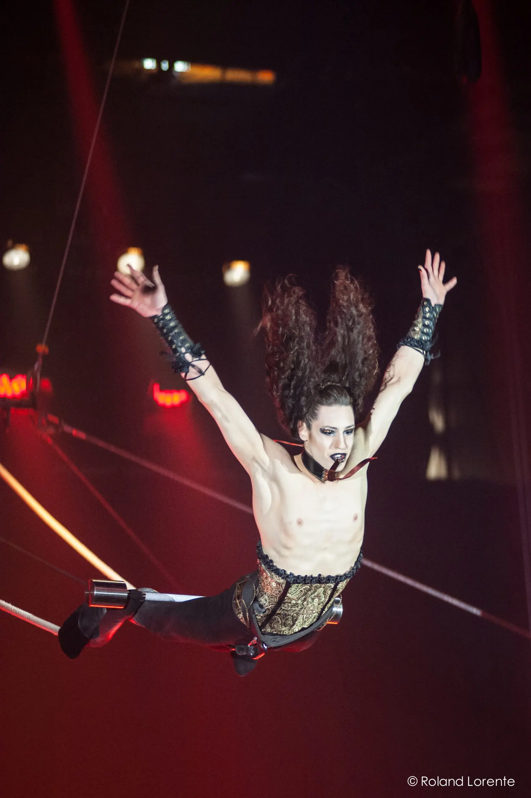 Une artiste de cirque aux cheveux longs, au maquillage spectaculaire et au corset métallique est suspendue dans les airs par des fils - un numéro à couper le souffle qui rappelle l'art de Célien Pinon - les bras tendus sur un fond sombre avec des lumières de scène rouges.