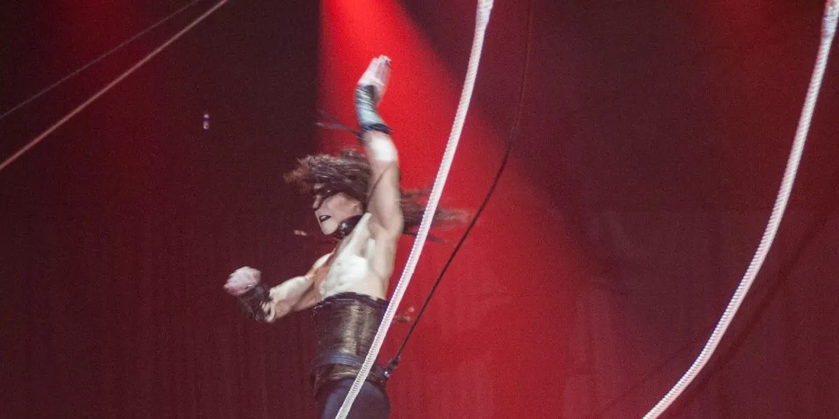 Célien Pinon se produit en plein vol sur un trapèze sous des lumières rouges spectaculaires, un bras levé et les cheveux en l'air, sur un fond de rideau rouge saisissant.