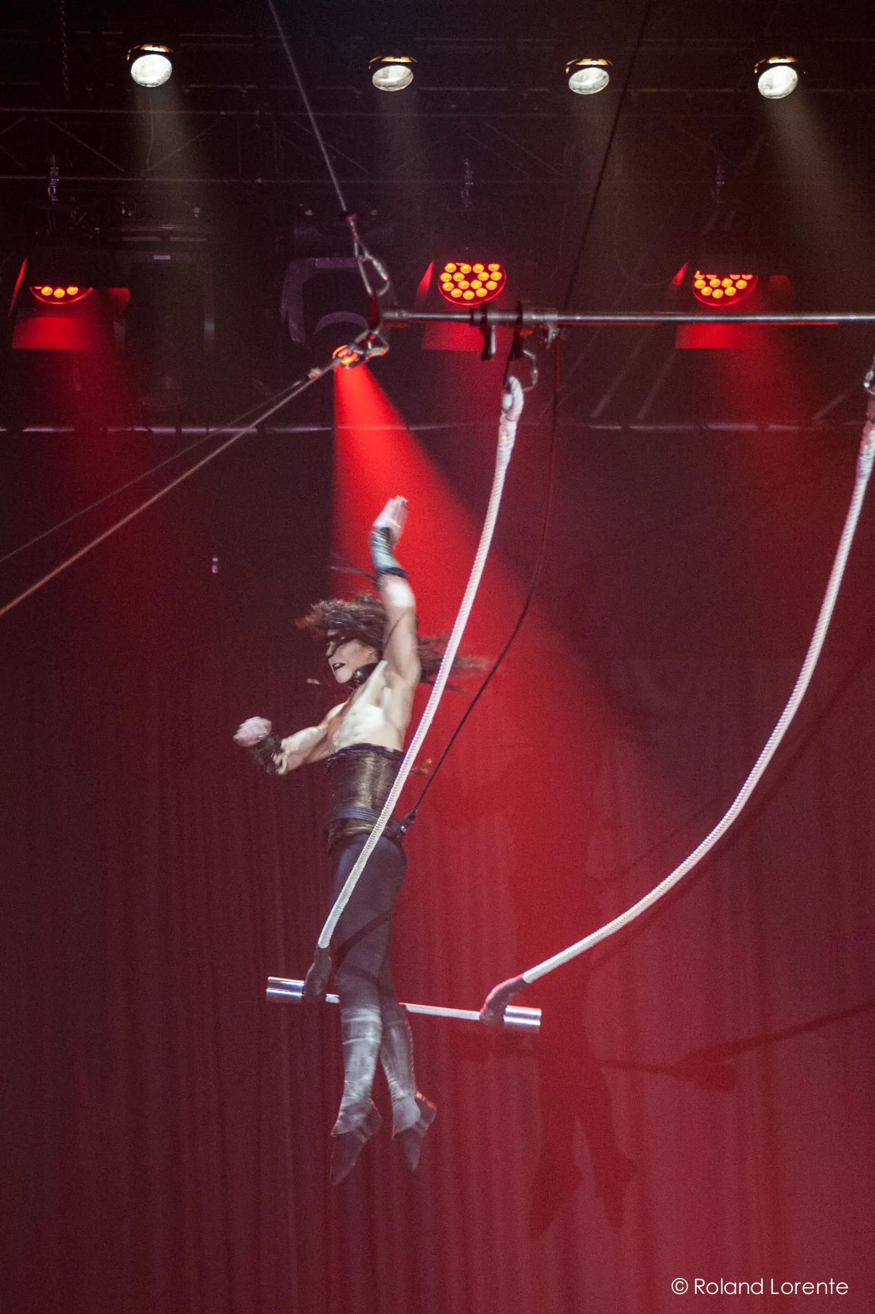 Célien Pinon se produit en plein vol sur un trapèze sous des lumières rouges spectaculaires, un bras levé et les cheveux en l'air, sur un fond de rideau rouge saisissant.