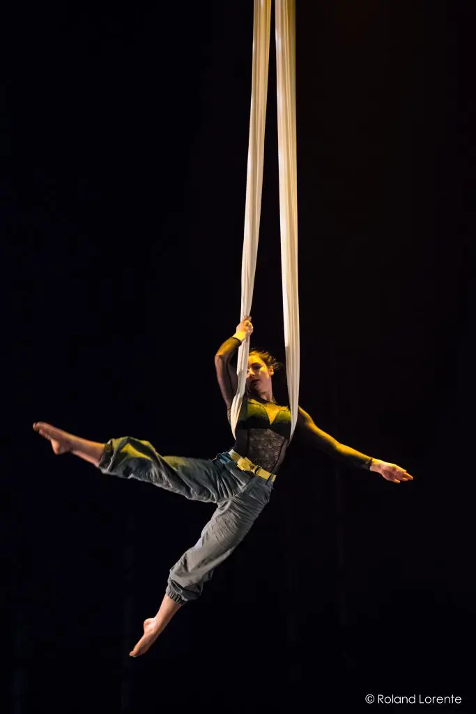 L'artiste aérienne Cindy Dumont est suspendue à des soies blanches, en équilibre avec une jambe tendue à l'horizontale et les bras agrippés au tissu. Elle porte un haut noir transparent et un pantalon gris sur un fond sombre.
