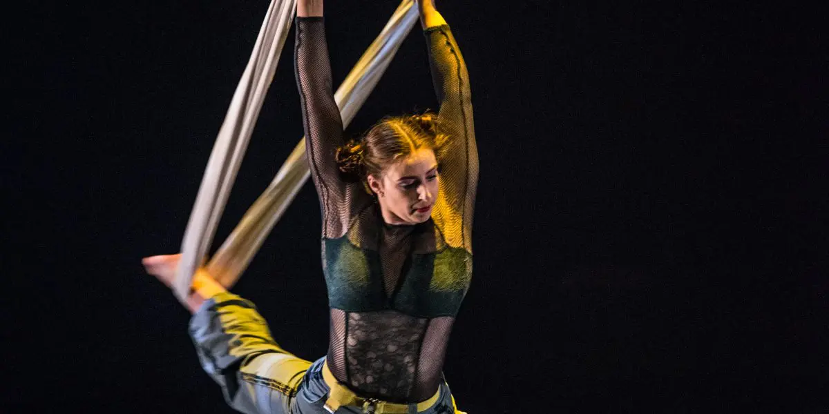 Une voltigeuse, Cindy Dumont, vêtue d'une tenue verte, exécute un grand écart en étant suspendue par des câbles aériens blancs au-dessus d'un canapé vert sur une scène faiblement éclairée.