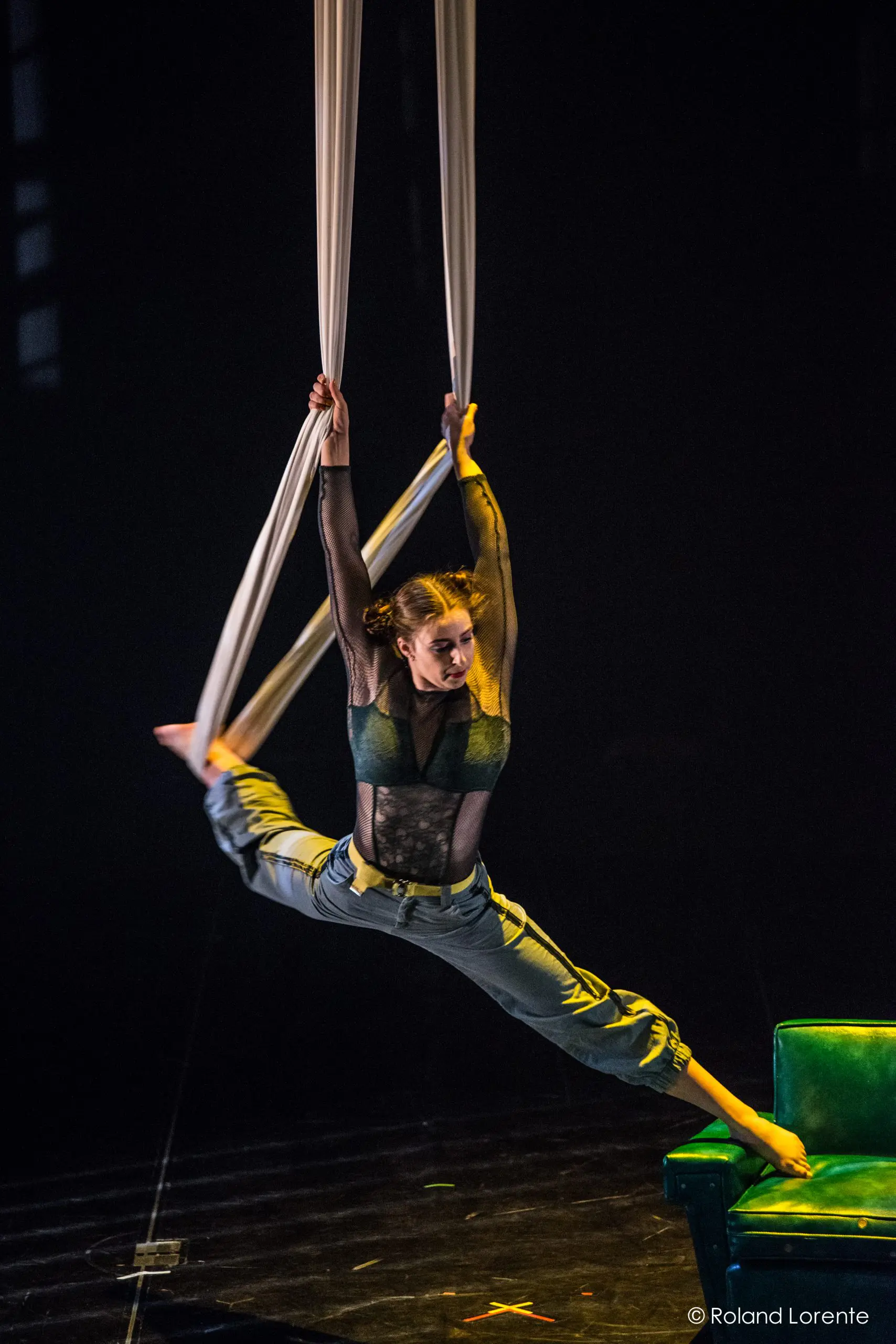 Une voltigeuse, Cindy Dumont, vêtue d'une tenue verte, exécute un grand écart en étant suspendue par des câbles aériens blancs au-dessus d'un canapé vert sur une scène faiblement éclairée.