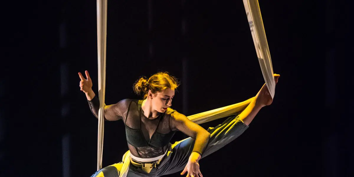 Une voltigeuse, Cindy Dumont, exécute une pose acrobatique en plein vol, se balançant gracieusement sur des soies aériennes blanches sur un fond sombre, avec un éclairage de scène dramatique mettant en valeur ses mouvements.
