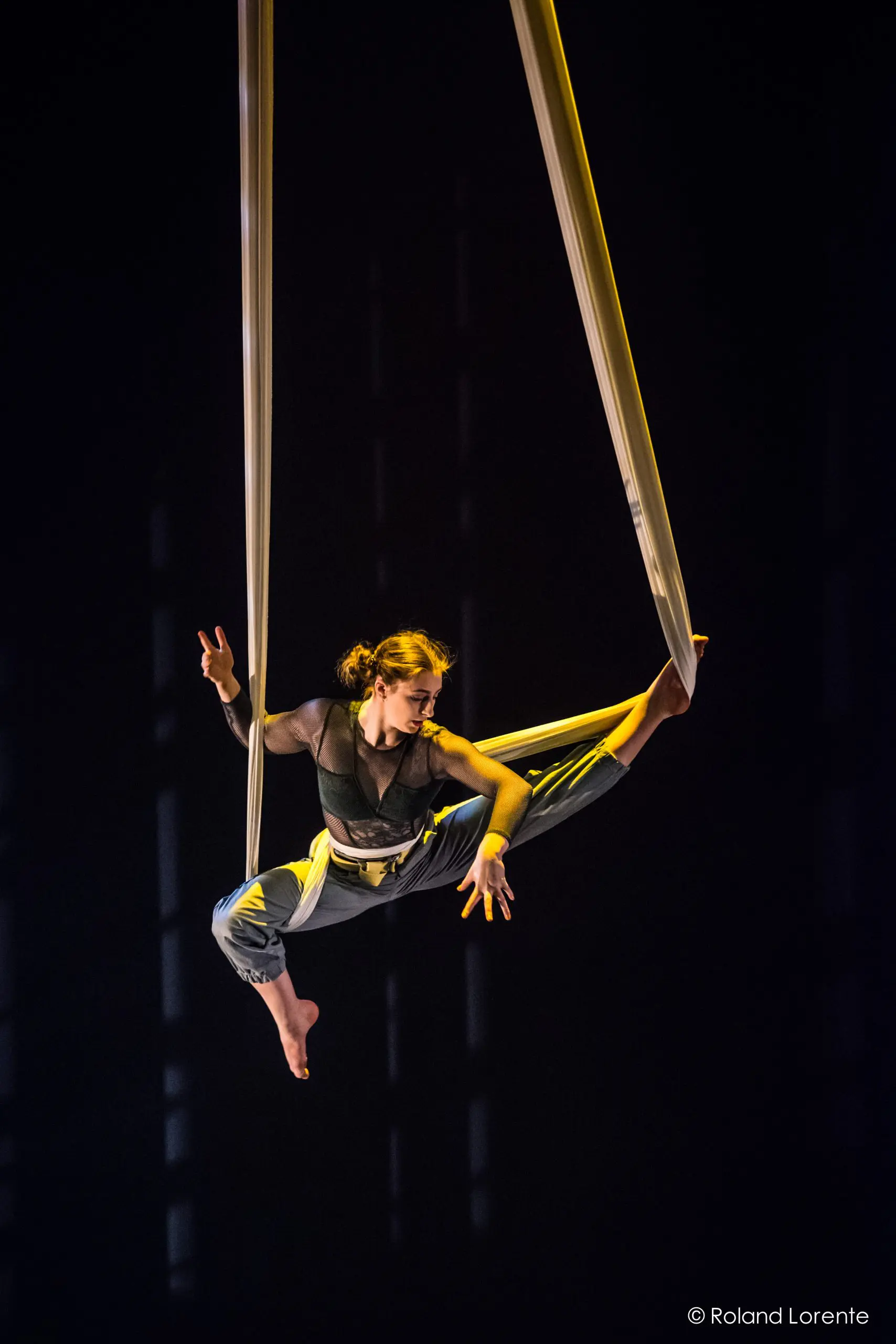 Une voltigeuse, Cindy Dumont, exécute une pose acrobatique en plein vol, se balançant gracieusement sur des soies aériennes blanches sur un fond sombre, avec un éclairage de scène dramatique mettant en valeur ses mouvements.