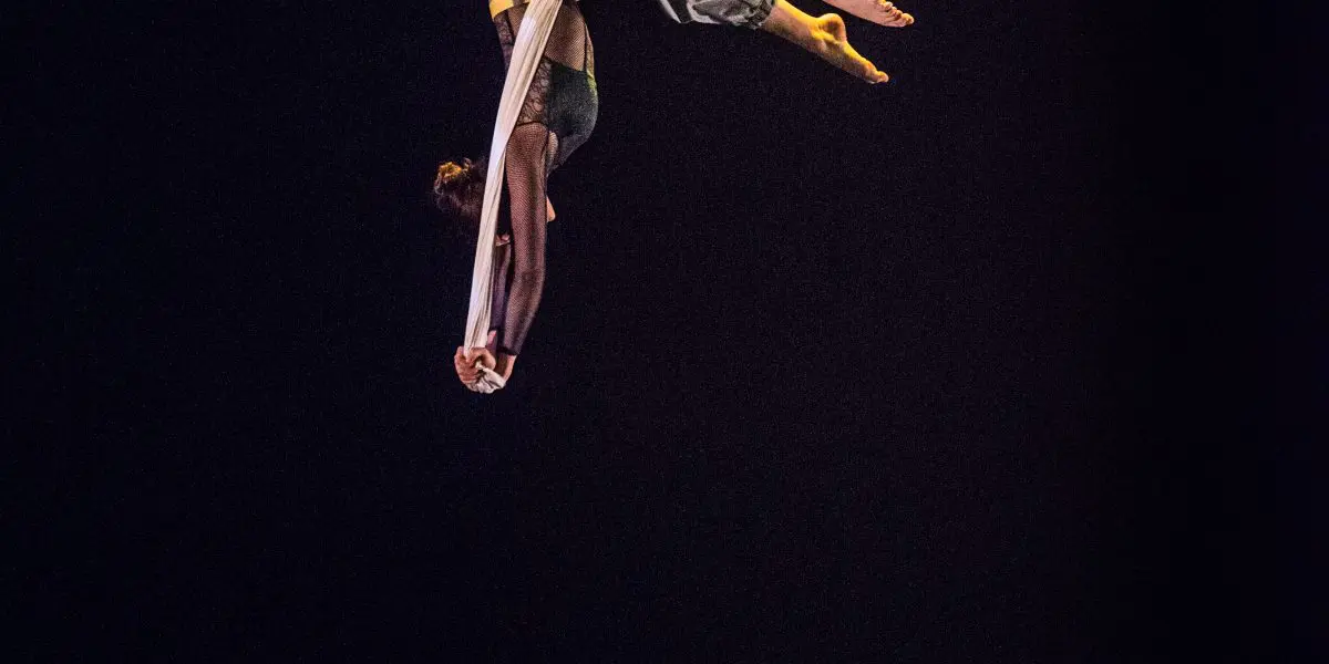 La voltigeuse Cindy Dumont effectue une pose inversée sur des soies blanches, suspendue dans les airs sur une scène sombre. Un fauteuil vert et un petit objet sont visibles en dessous, tandis qu'un éclairage dramatique met en valeur l'artiste.