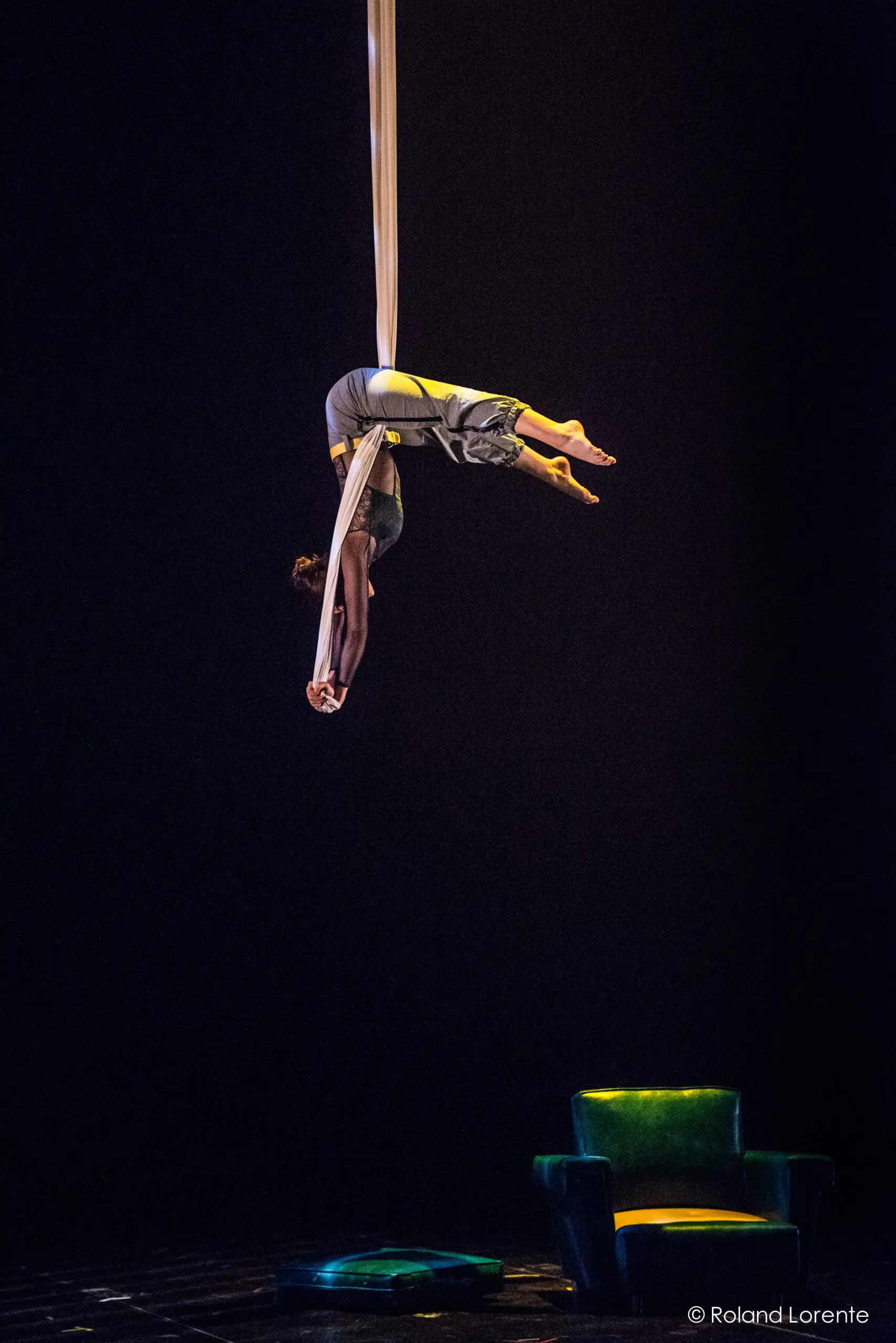 La voltigeuse Cindy Dumont effectue une pose inversée sur des soies blanches, suspendue dans les airs sur une scène sombre. Un fauteuil vert et un petit objet sont visibles en dessous, tandis qu'un éclairage dramatique met en valeur l'artiste.