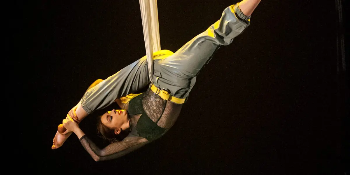 L'artiste aérienne Cindy Dumont, suspendue à l'envers dans des soies blanches, les jambes repliées au-dessus de sa tête, pose à côté d'un fauteuil vert sur une scène sombre sous un éclairage dramatique.