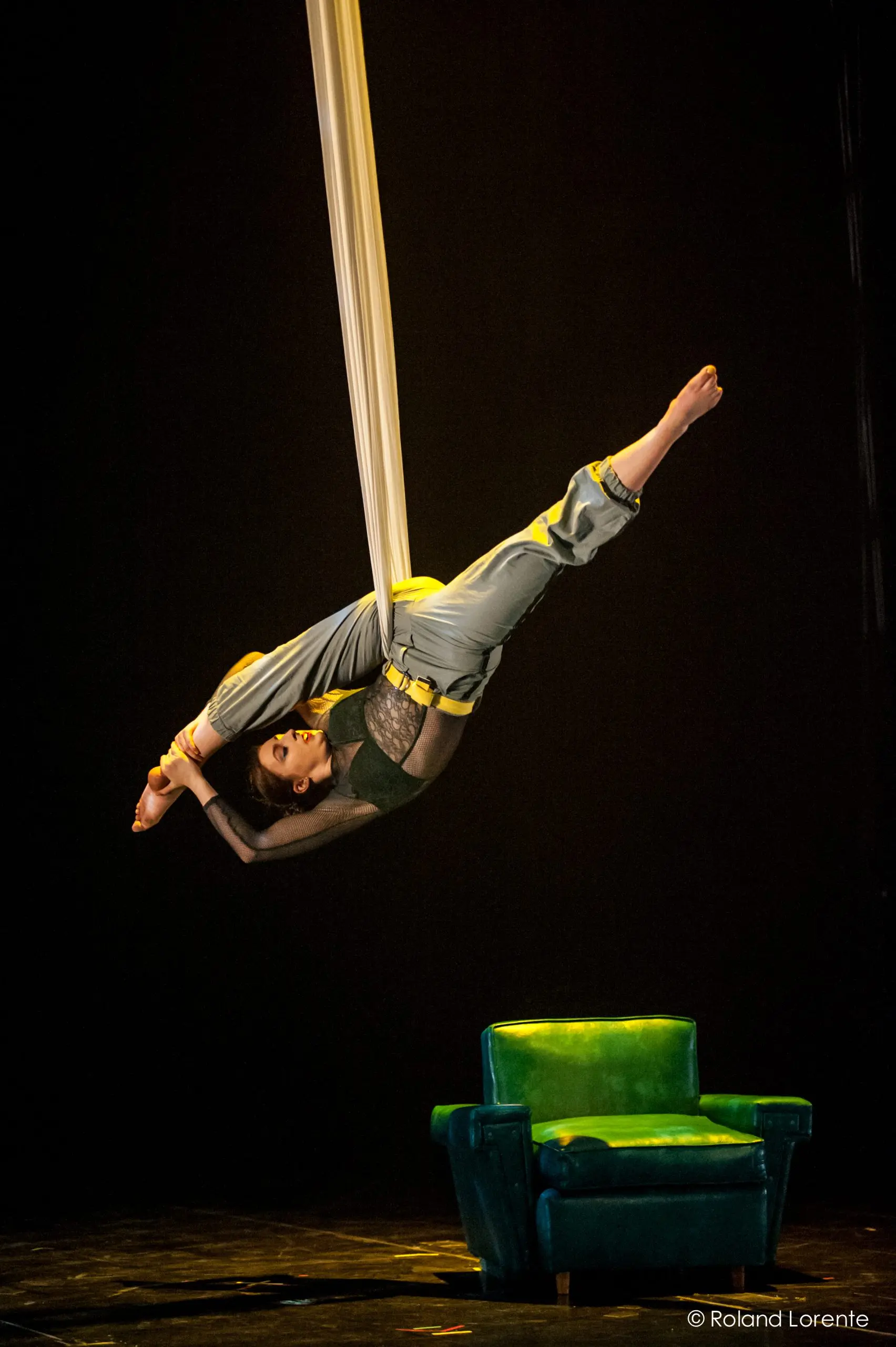 L'artiste aérienne Cindy Dumont, suspendue à l'envers dans des soies blanches, les jambes repliées au-dessus de sa tête, pose à côté d'un fauteuil vert sur une scène sombre sous un éclairage dramatique.
