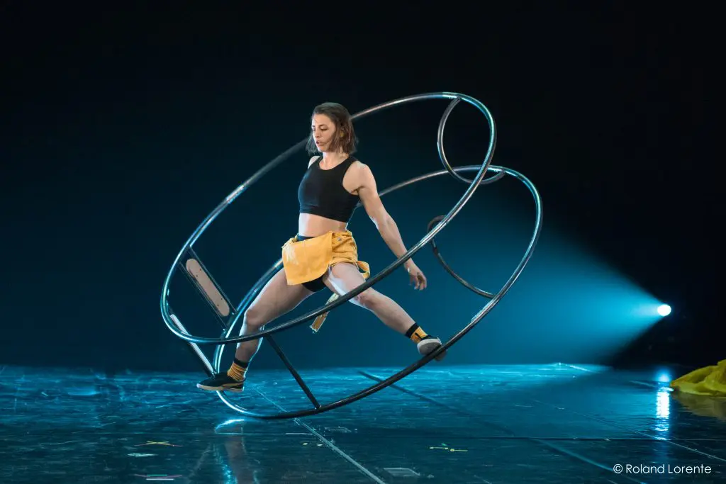 Clara Prezzavento, vêtue d'un haut noir et d'un short jaune, se tient habilement en équilibre à l'intérieur d'une grande roue Cyr en métal sur une scène faiblement éclairée.