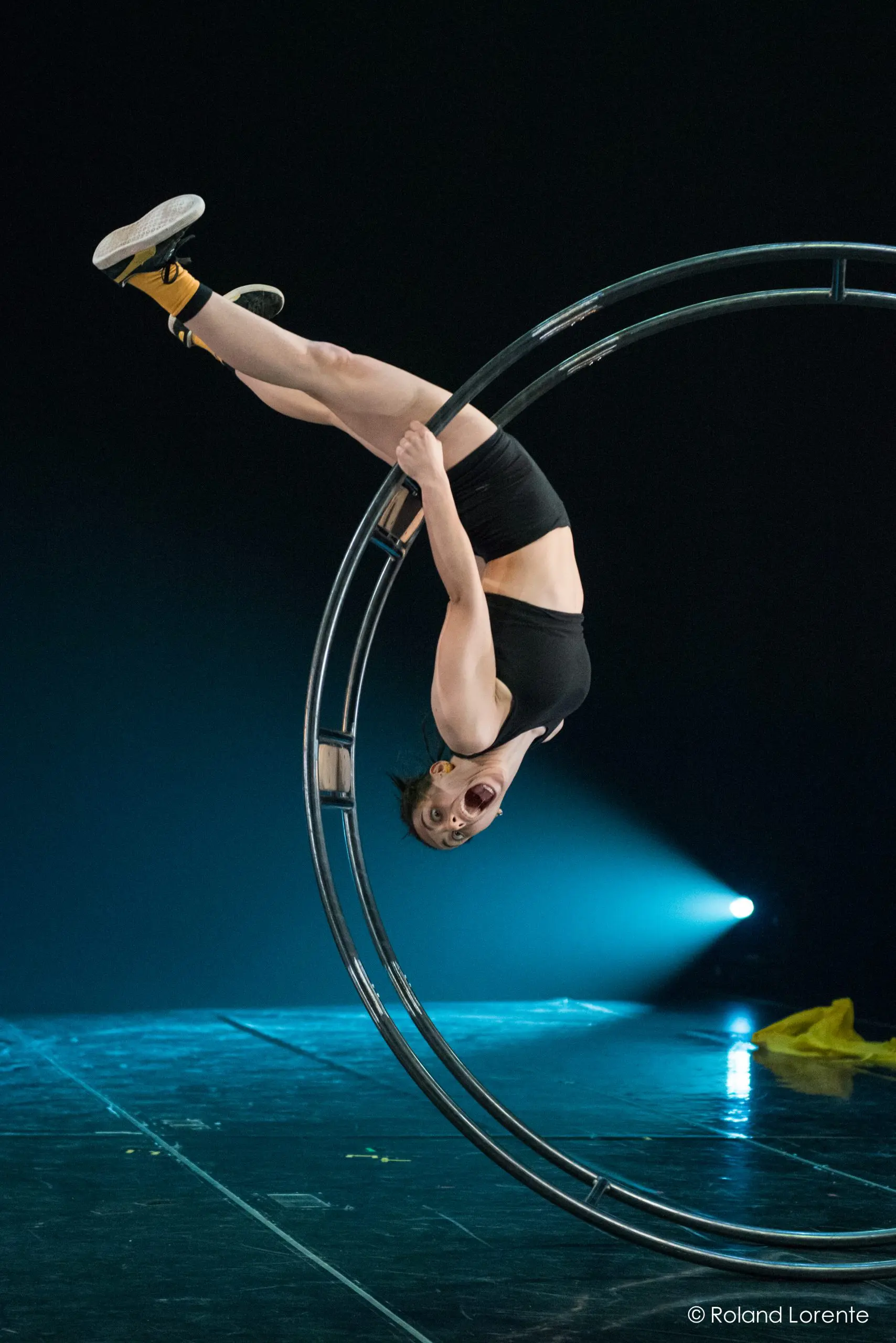 Clara Prezzavento, vêtue d'une tenue de sport noire et de chaussettes jaunes, est suspendue la tête en bas à un grand cerceau métallique sur la scène, éclairée par un projecteur et un tissu jaune posé sur le sol.