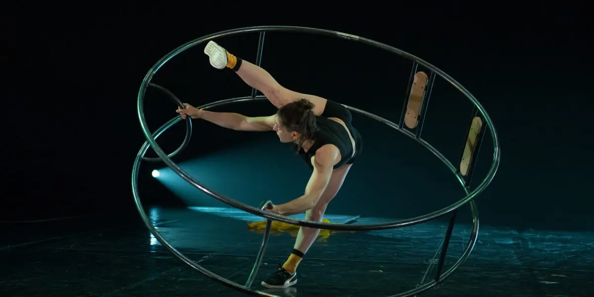 Clara Prezzavento se tient en équilibre à l'intérieur d'une grande roue métallique, étendant une jambe vers le haut tout en saisissant la jante à deux mains, sur une scène faiblement éclairée.