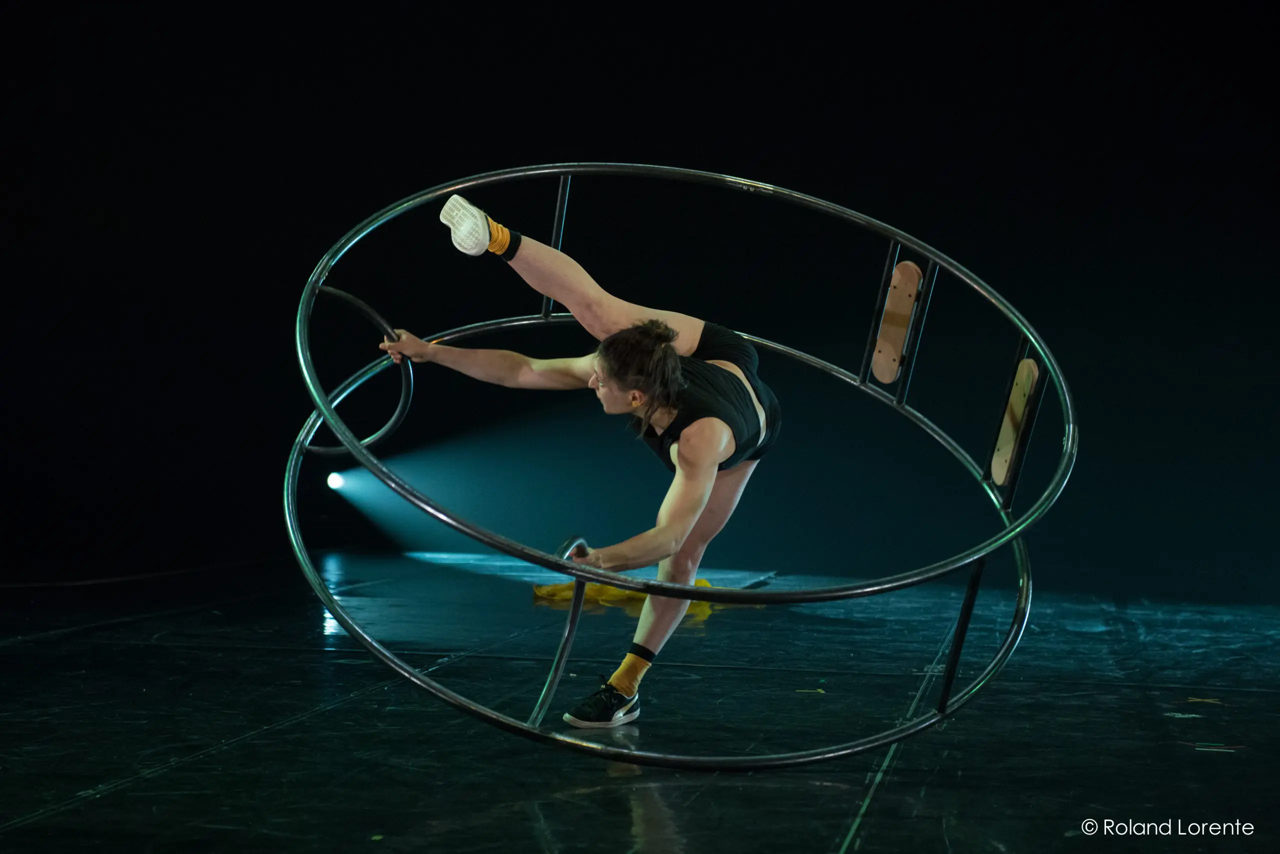 Clara Prezzavento se tient en équilibre à l'intérieur d'une grande roue métallique, étendant une jambe vers le haut tout en saisissant la jante à deux mains, sur une scène faiblement éclairée.