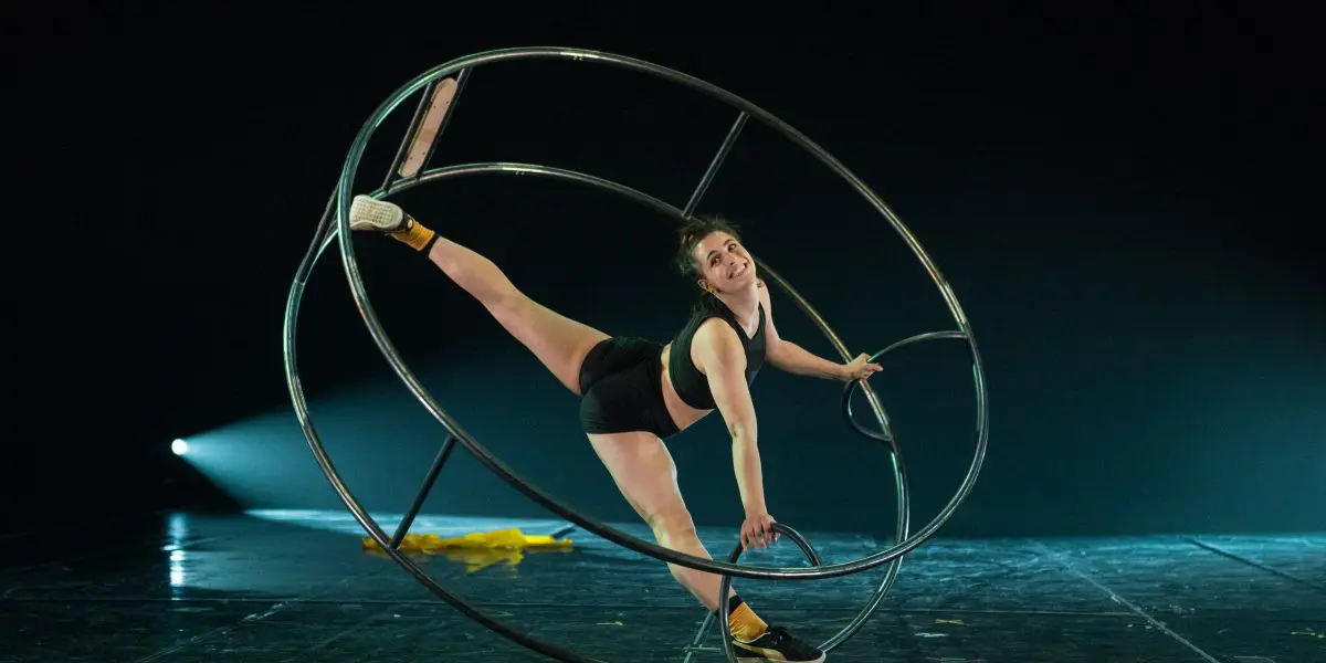 Clara Prezzavento, en tenue de sport, se tient en équilibre à l'intérieur d'une grande roue métallique sur la scène, étendant une jambe vers le haut tout en s'agrippant au cadre. La scène est faiblement éclairée par un projecteur en arrière-plan.