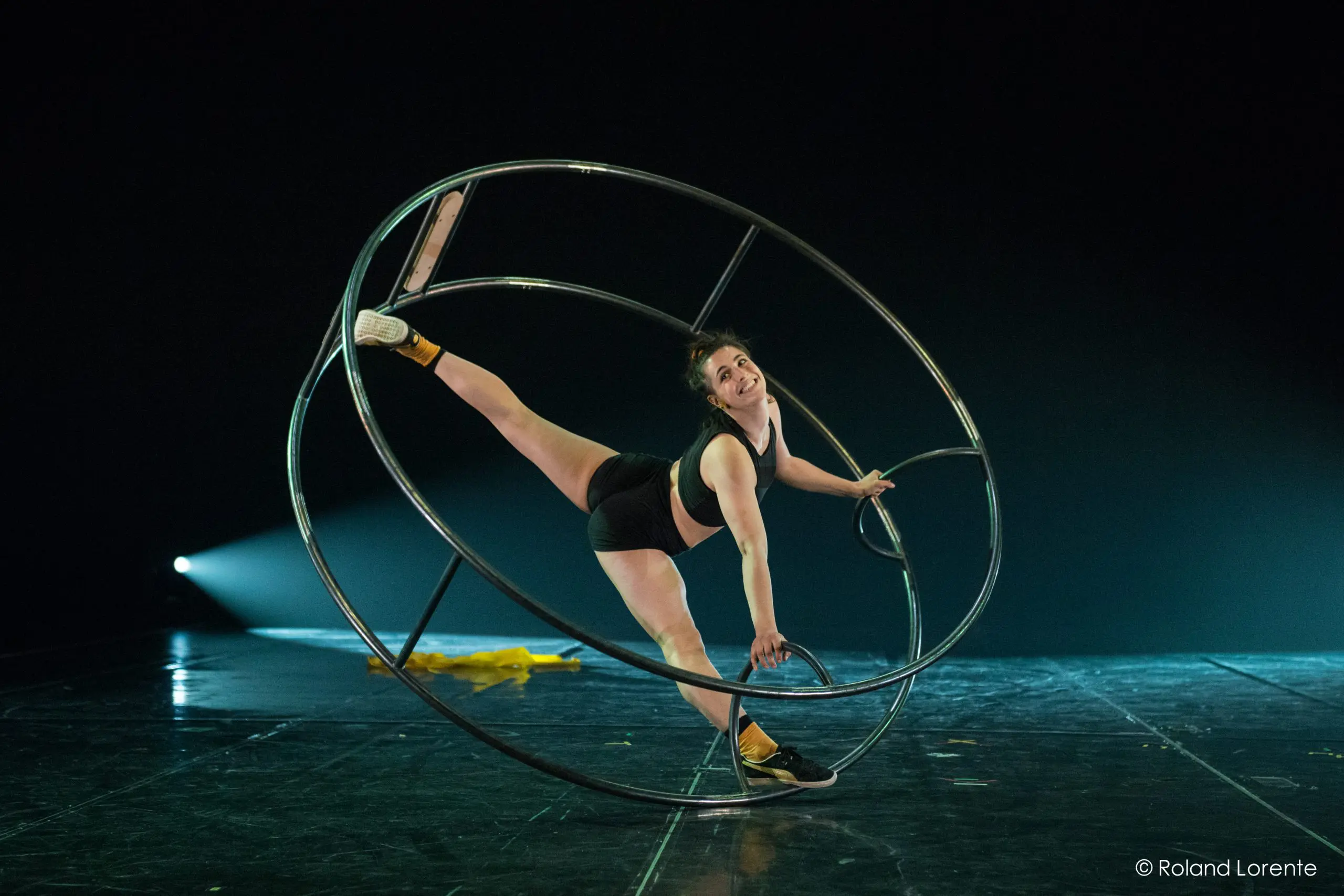 Clara Prezzavento, en tenue de sport, se tient en équilibre à l'intérieur d'une grande roue métallique sur la scène, étendant une jambe vers le haut tout en s'agrippant au cadre. La scène est faiblement éclairée par un projecteur en arrière-plan.