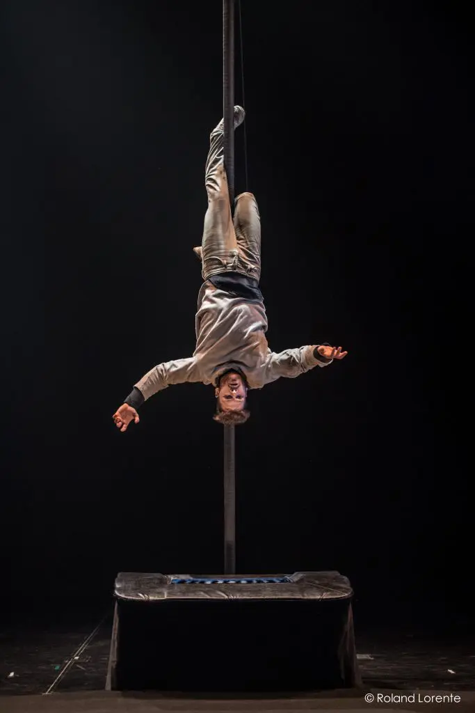 Raphaël Filiatreault est suspendu à un poteau vertical, en équilibre avec une jambe crochetée, les bras tendus au-dessus d'une plate-forme rembourrée sur une scène sombre.