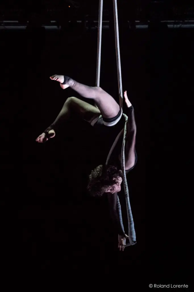 L'artiste aérienne Georgia Simpson, vêtue de noir, est suspendue à l'envers sur des soies aériennes sur un fond sombre, un bras tendu gracieusement vers le bas et les jambes pliées élégamment autour du tissu fluide.