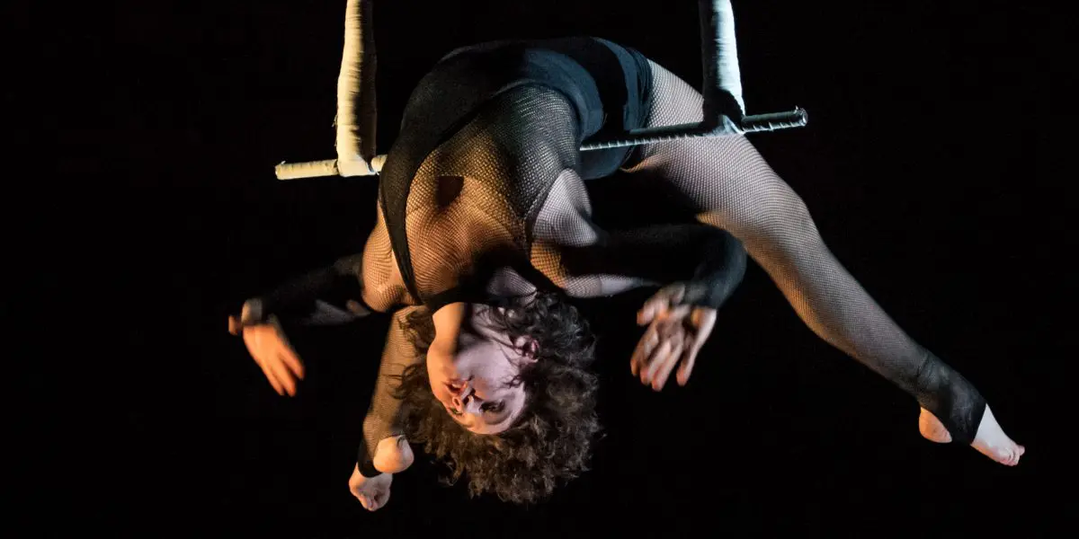 L'artiste aérienne Georgia Simpson effectue un saut à l'envers sur un trapèze, les bras étendus et la tête inclinée vers l'arrière, sur un fond sombre.