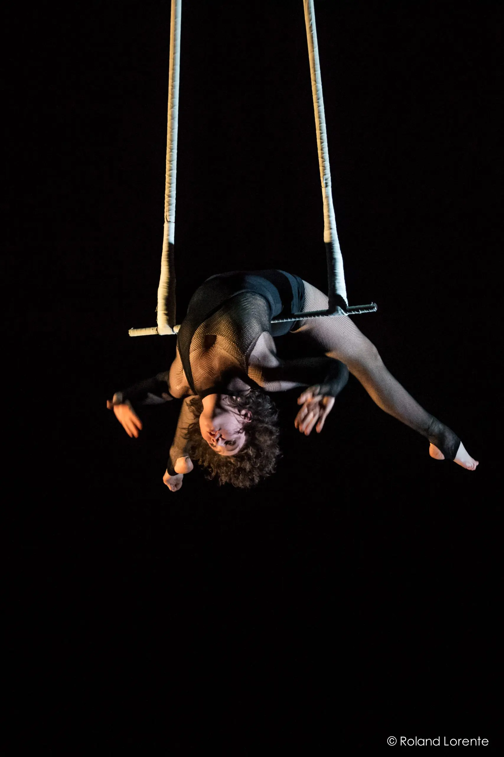 L'artiste aérienne Georgia Simpson effectue un saut à l'envers sur un trapèze, les bras étendus et la tête inclinée vers l'arrière, sur un fond sombre.