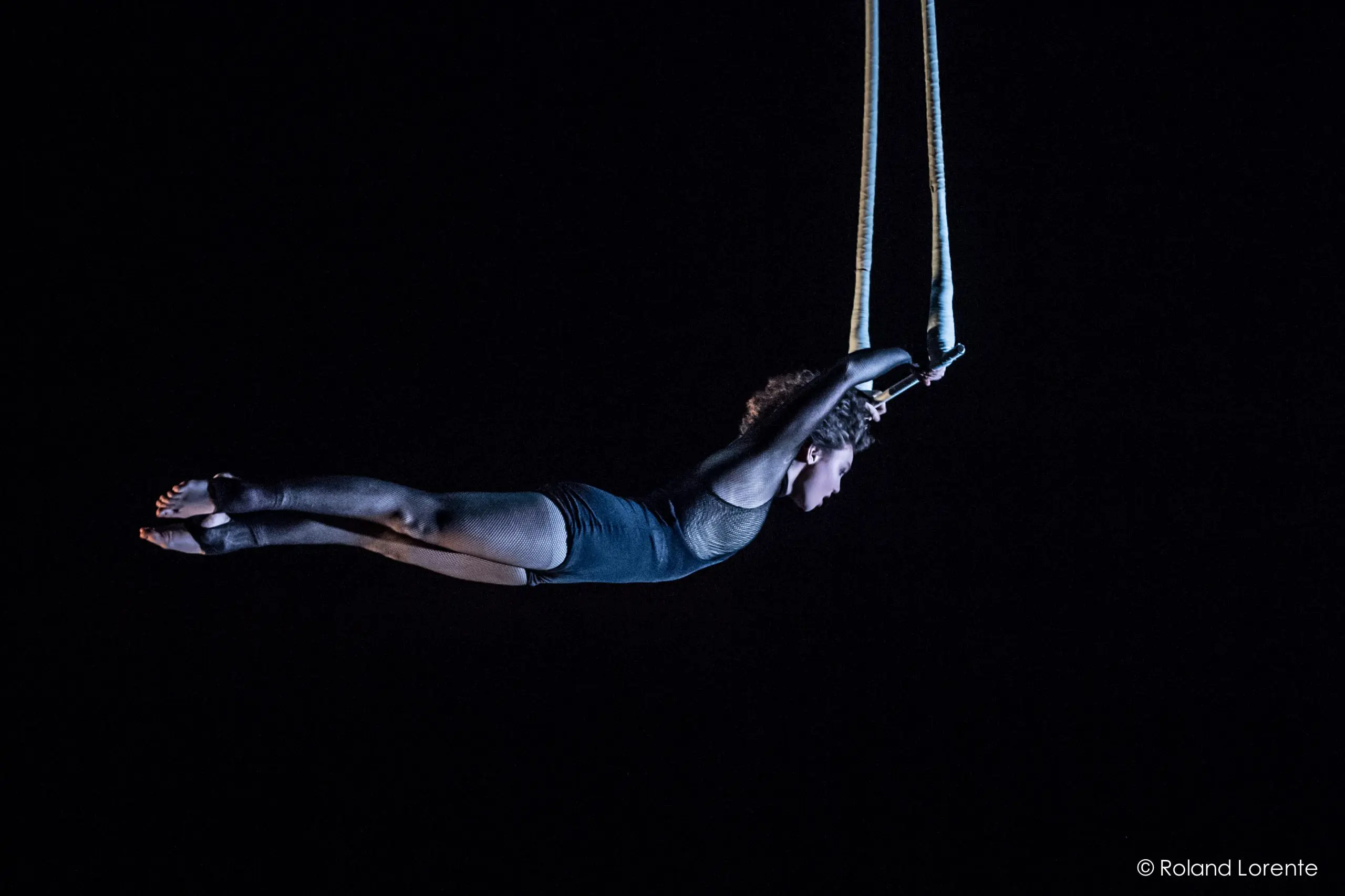 L'acrobate Georgia Simpson, vêtue d'un justaucorps sombre, évolue dans les airs sur des sangles suspendues sur un fond noir, saisissant les sangles à deux mains alors qu'elle étend son corps à l'horizontale.